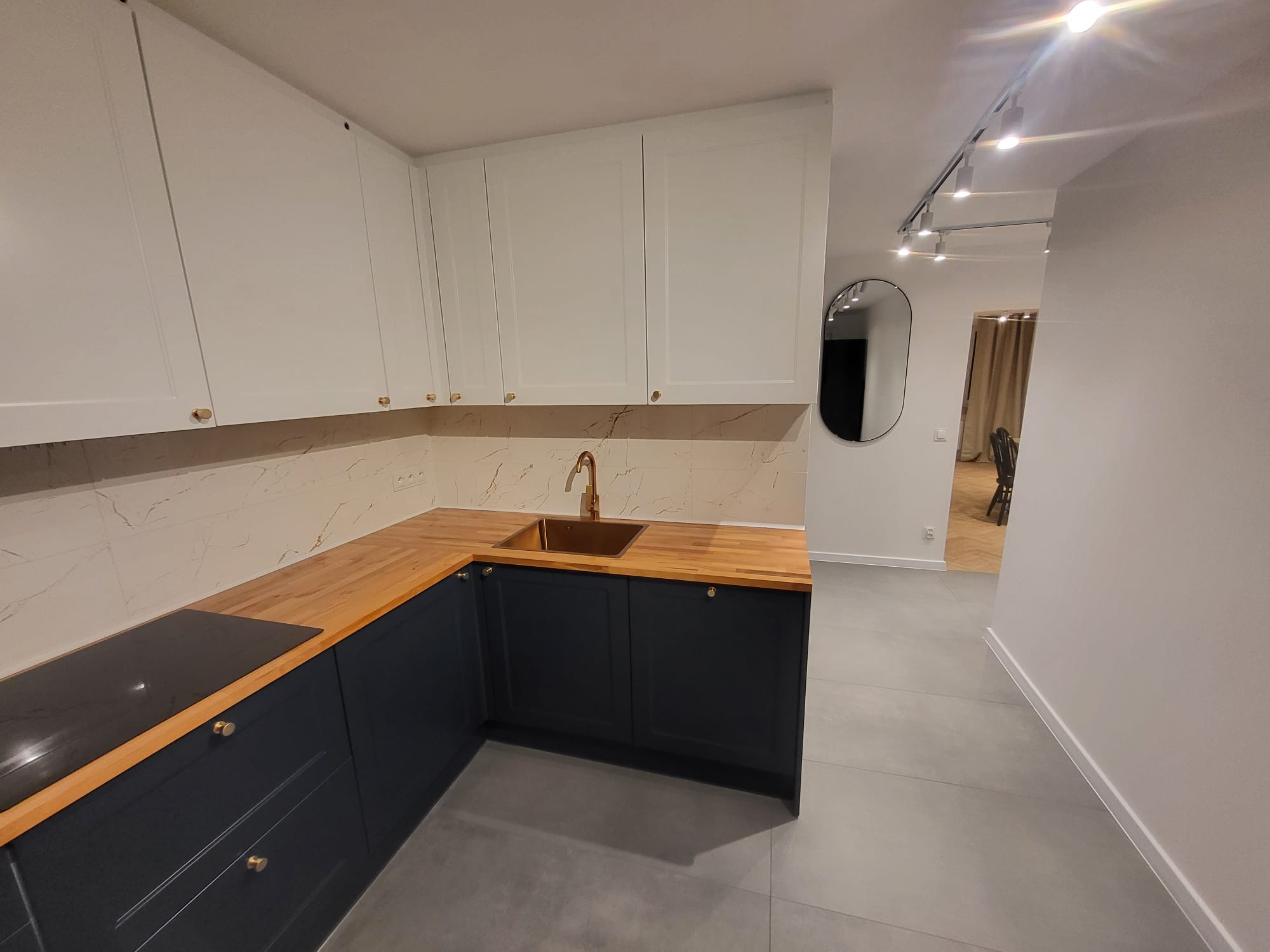 Wykończona kuchnia w bieli i granacie: drewniany blat, złota armatura, marmurowe płytki. Widok na salon z owalnym lustrem. Nowoczesne oświetlenie.