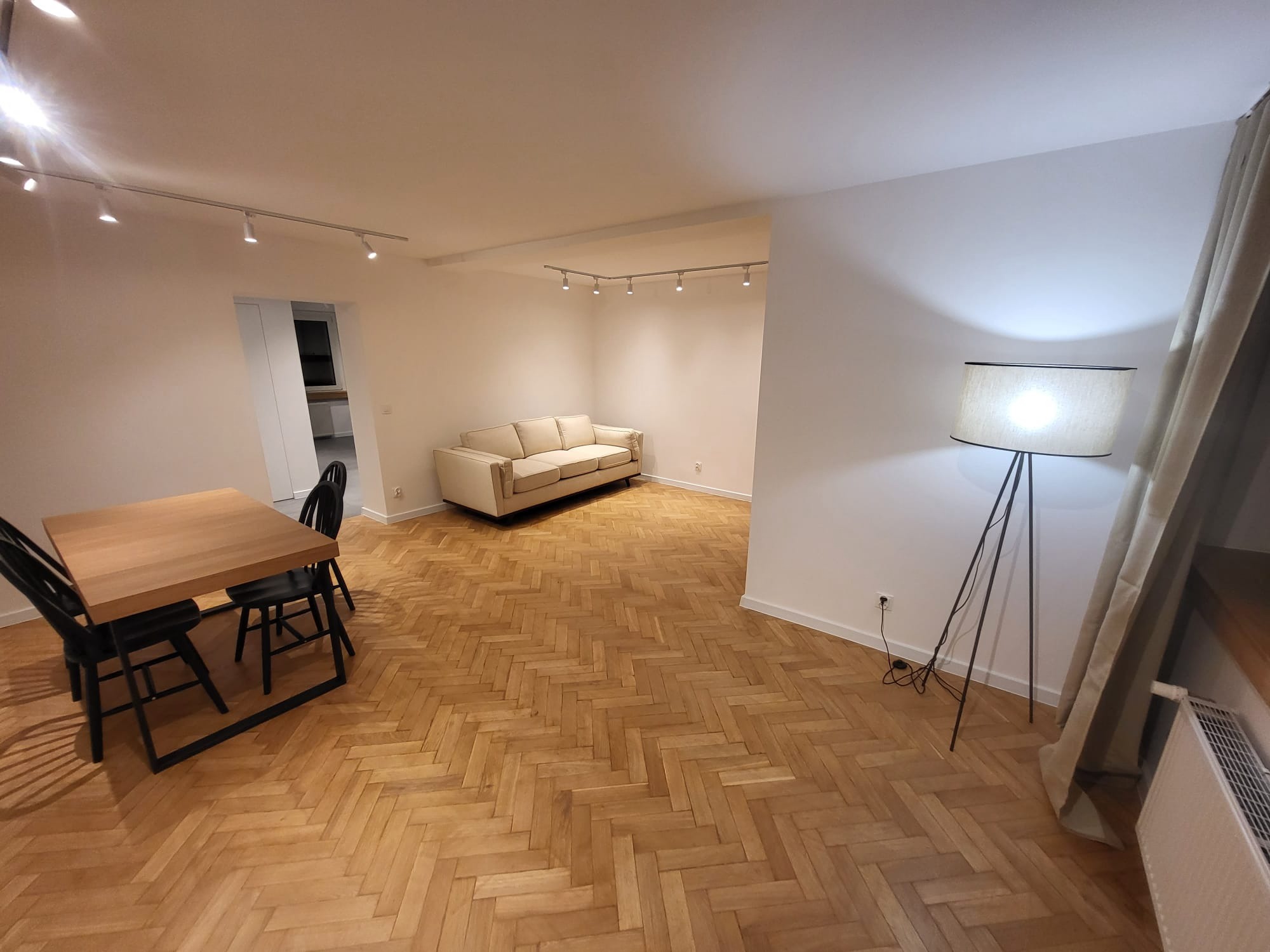 Jasne, przestronne wnętrze po remoncie z parkietem w jodełkę, nowoczesnym oświetleniem, sofą i stołem z krzesłami. Widok na odnowione mieszkanie.