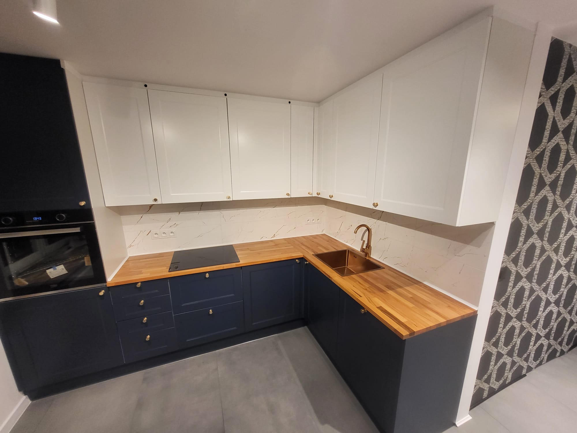 Nowoczesna kuchnia z drewnianym blatem i białymi szafkami górnymi. Widoczny złoty kran i marmurowe wykończenie ściany. Ciemne szafki dolne z złotymi uchwytami.