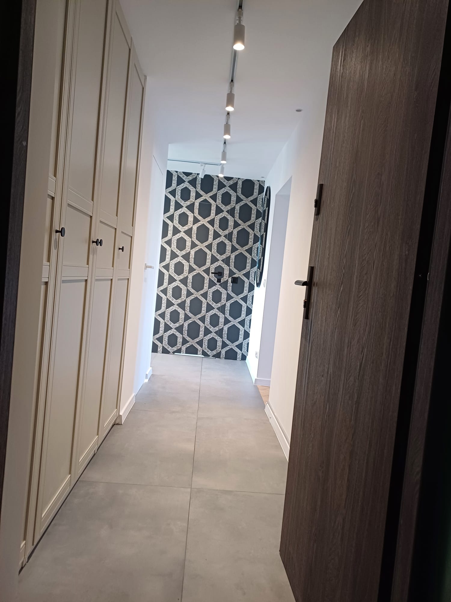 Nowoczesny korytarz z szafą w zabudowie, szarą podłogą i wzorzystą tapetą. Otwarta drewniana drzwi dodają głębi. Oświetlenie punktowe.