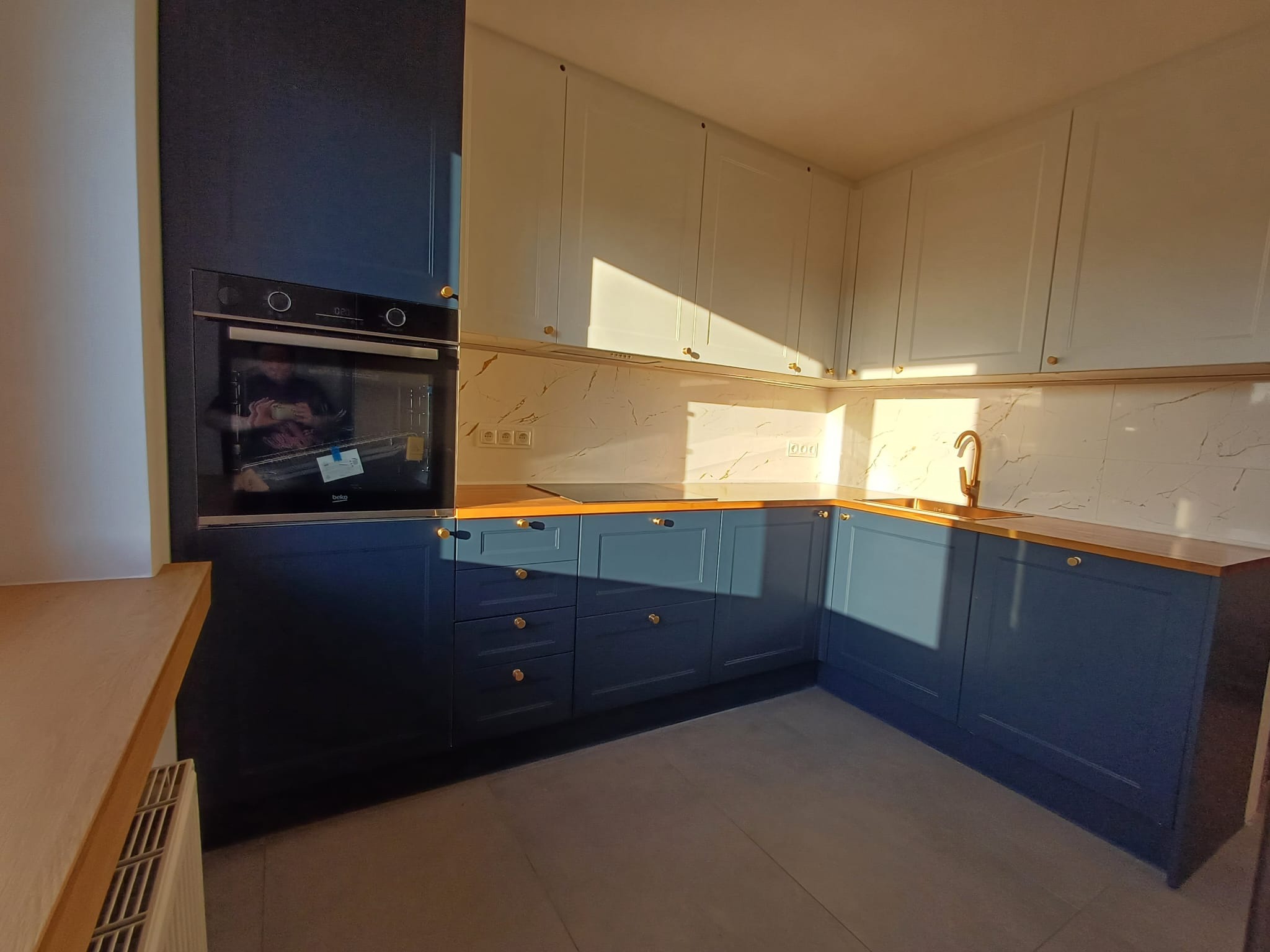 Nowoczesna, narożna kuchnia w odcieniach bieli i granatu, z drewnianym blatem i złotymi akcentami. Widoczne szafki górne i dolne, piekarnik oraz marmurowa ściana.