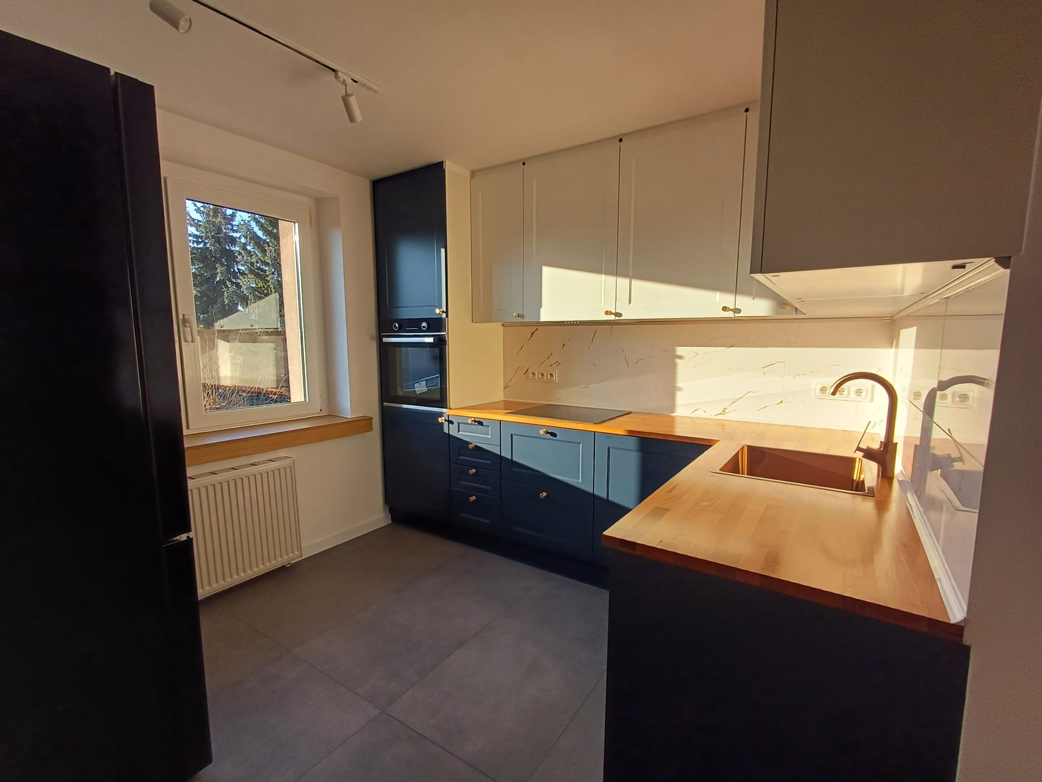 Nowoczesna kuchnia z granatowymi szafkami, drewnianym blatem i marmurowym backsplashiem w promieniach słońca. Widok na okno i grzejnik.
