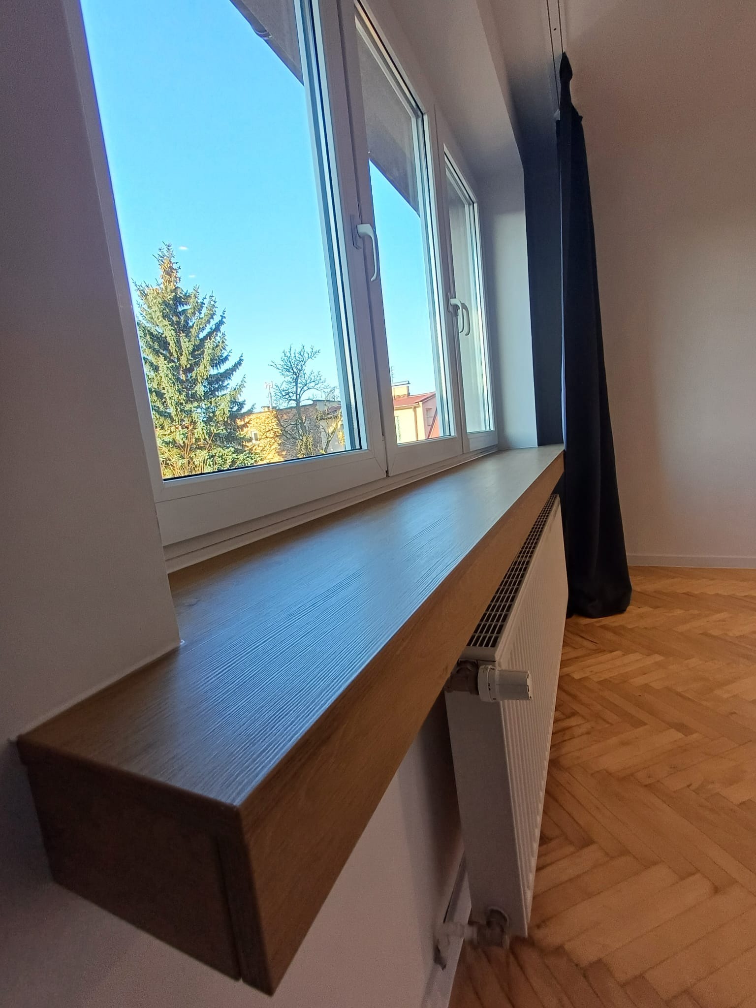 Zamontowany drewniany parapet w Żorach z widokiem na zewnątrz. Grzejnik pod parapetem, parkiet na podłodze i ciemne zasłony w tle.