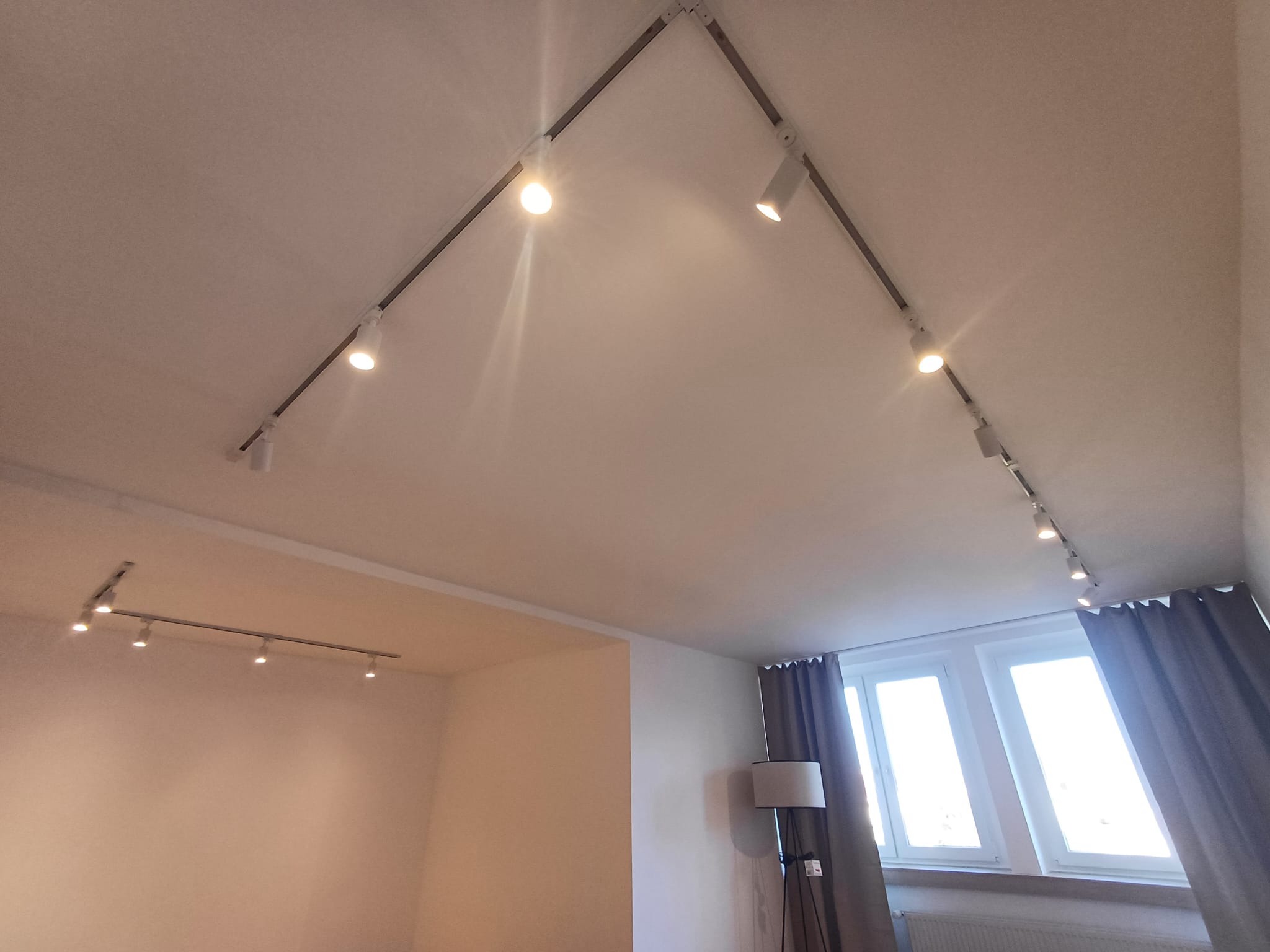 Biały sufit z zamontowanym systemem szynoprzewodów z reflektorami, oświetlającymi wnętrze. Widoczne okno z zasłonami i lampa podłogowa.