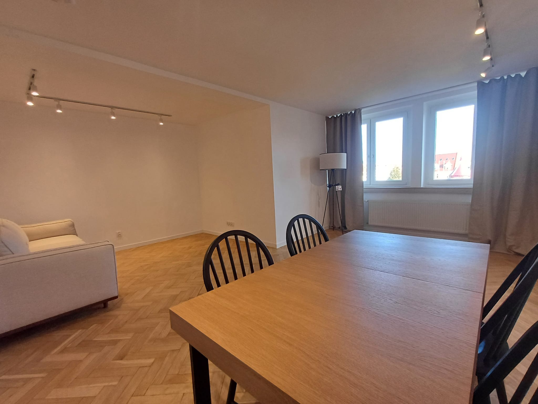 Jasne wnętrze po remoncie: parkiet w jodełkę, sofa, drewniany stół z krzesłami, lampa podłogowa, okna z zasłonami, oświetlenie sufitowe. Widok na pokój dzienny.