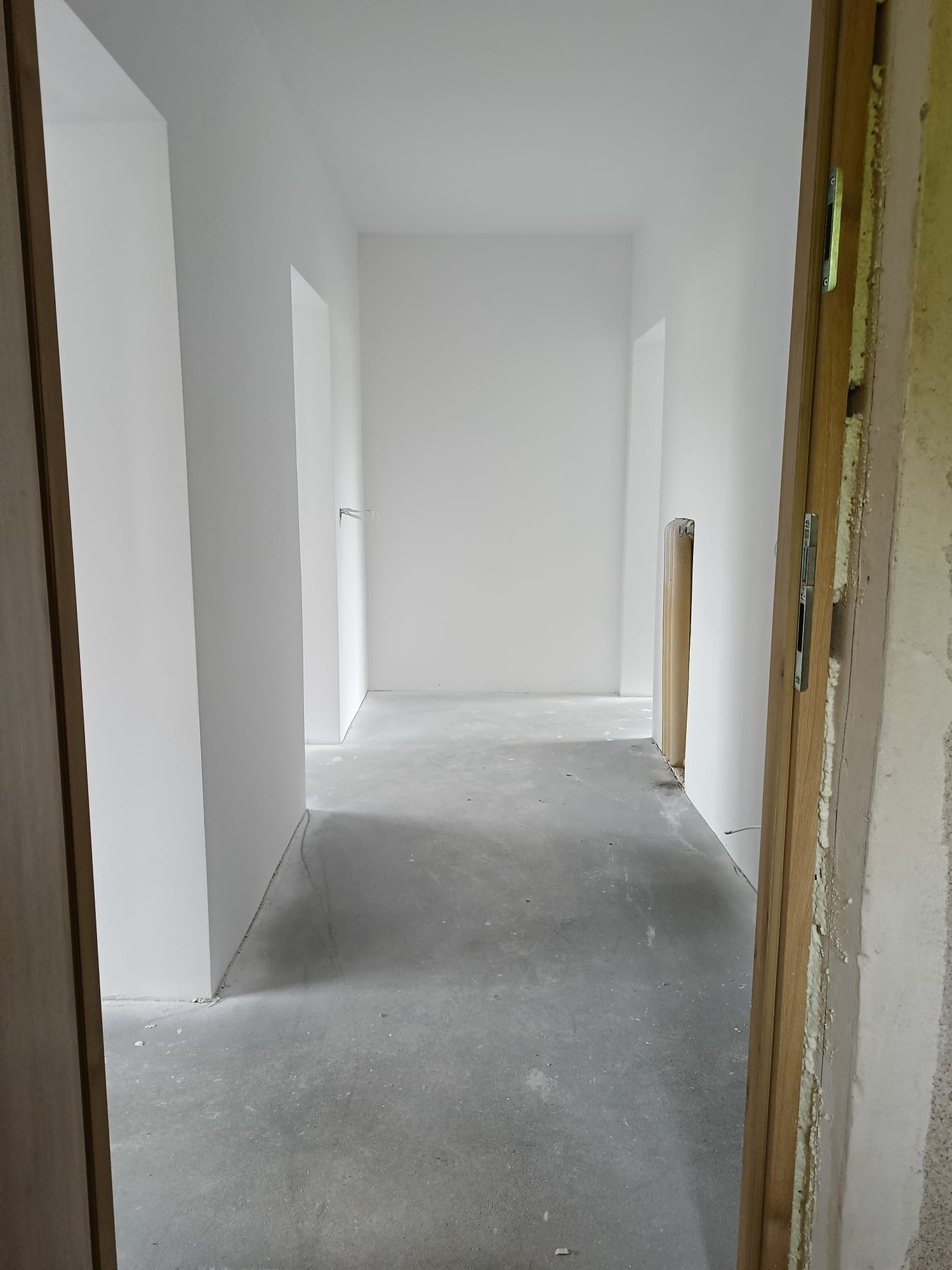 Wnętrze w trakcie prac wykończeniowych: białe ściany, surowa podłoga, widoczne ościeżnice. Fragment otwartych drzwi z prawej strony kadru.