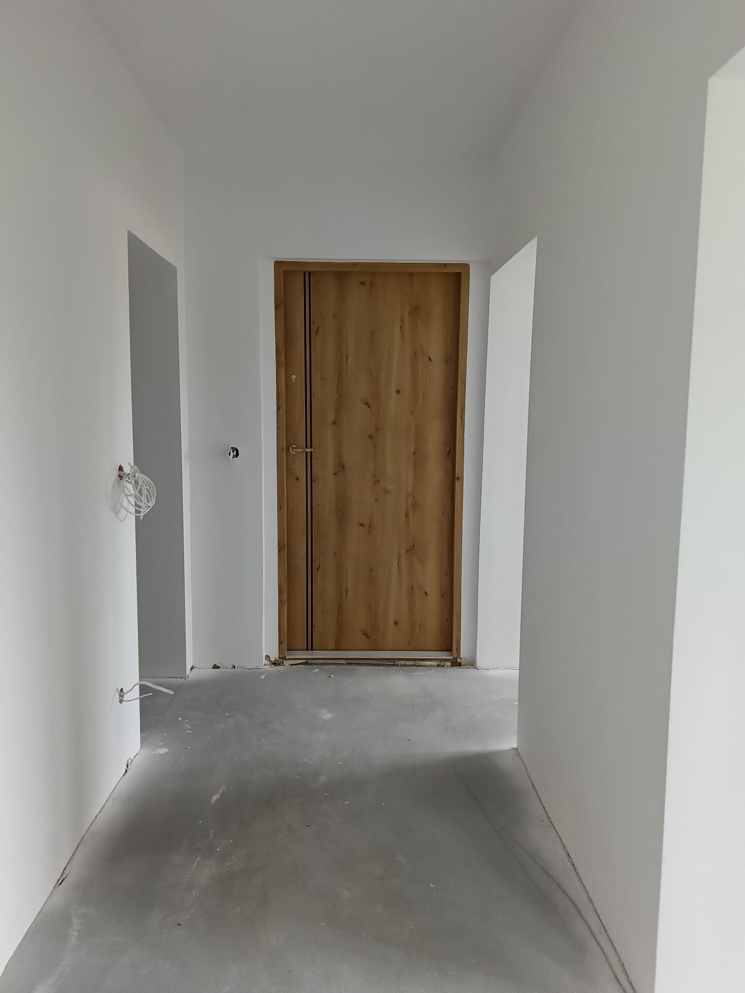 Wnętrze po remoncie: drewniane drzwi z czarnymi paskami na tle białych ścian i szarej posadzki. Widoczne niedociągnięcia wykończeniowe, gniazdka bez osprzętu.