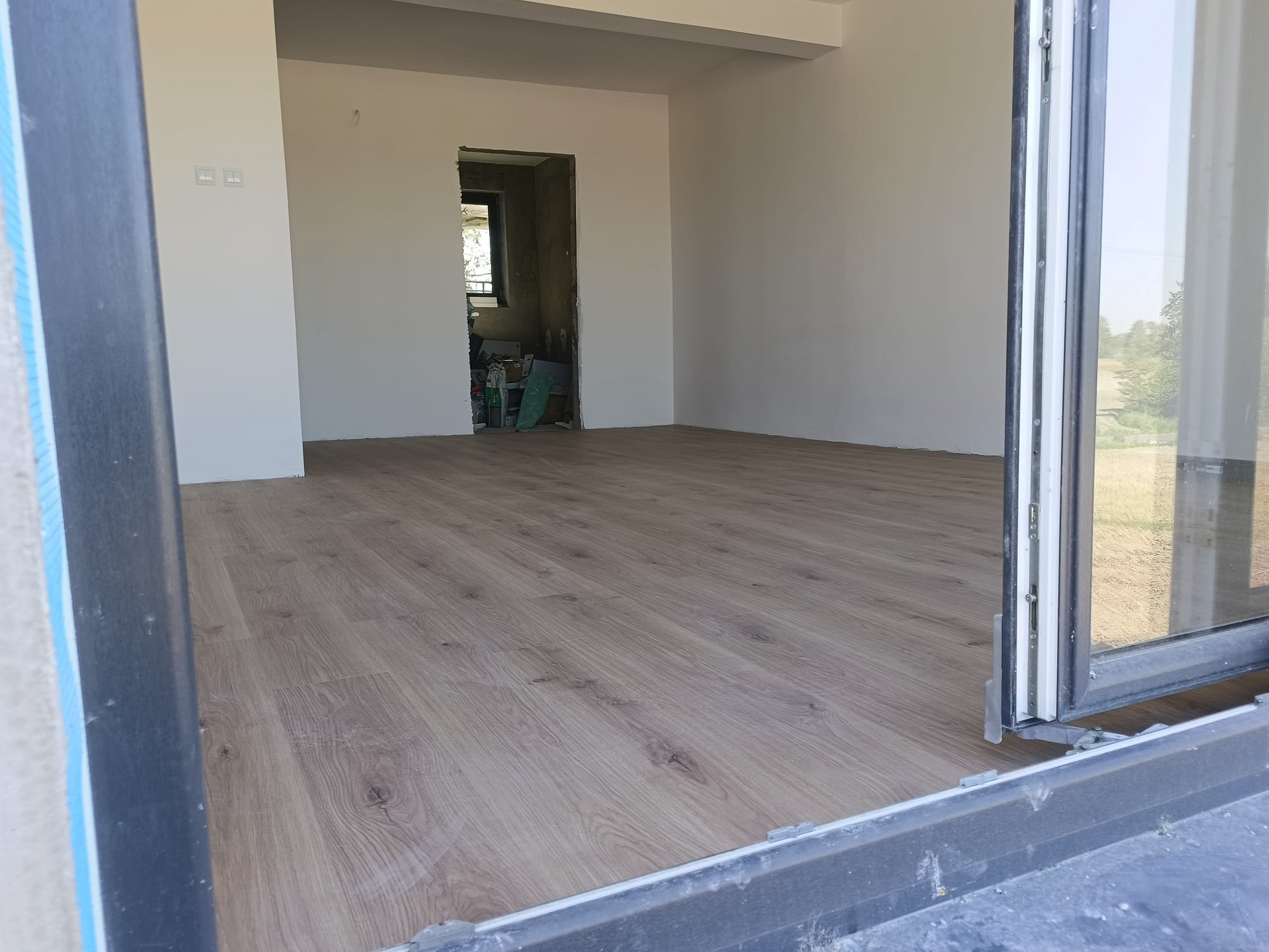 Jasne panele podłogowe ułożone w pustym pokoju, widok zza otwartych drzwi balkonowych. Białe ściany, w tle widoczne drzwi do innego pomieszczenia.