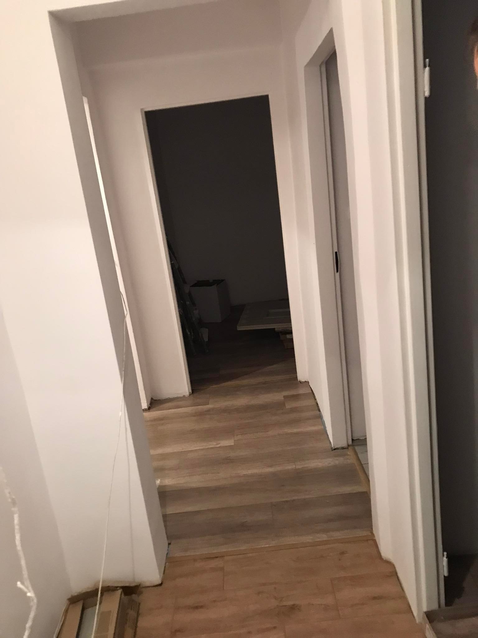 Korytarz z nowo ułożonymi panelami podłogowymi w odcieniach drewna, widok na otwarte drzwi do kolejnego pomieszczenia, ściany pomalowane na biało, widoczne niedociągnięcia w wykończeniu przy podłodze.
