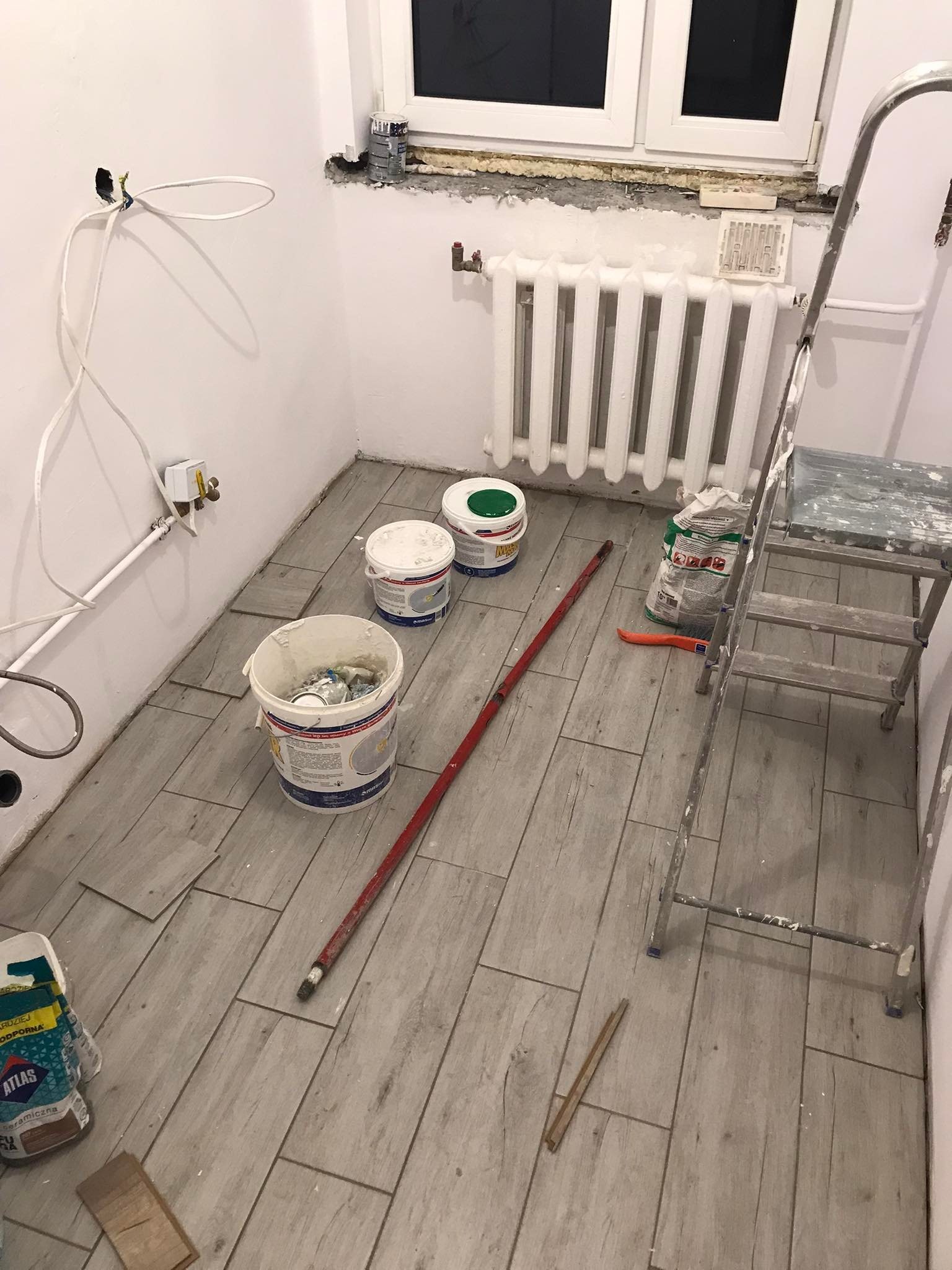 Wnętrze łazienki w trakcie remontu: częściowo ułożona podłoga z paneli imitujących drewno, drabina malarska, wiadra z farbą, worek z klejem Atlas, biały grzejnik pod oknem, wystające kable...