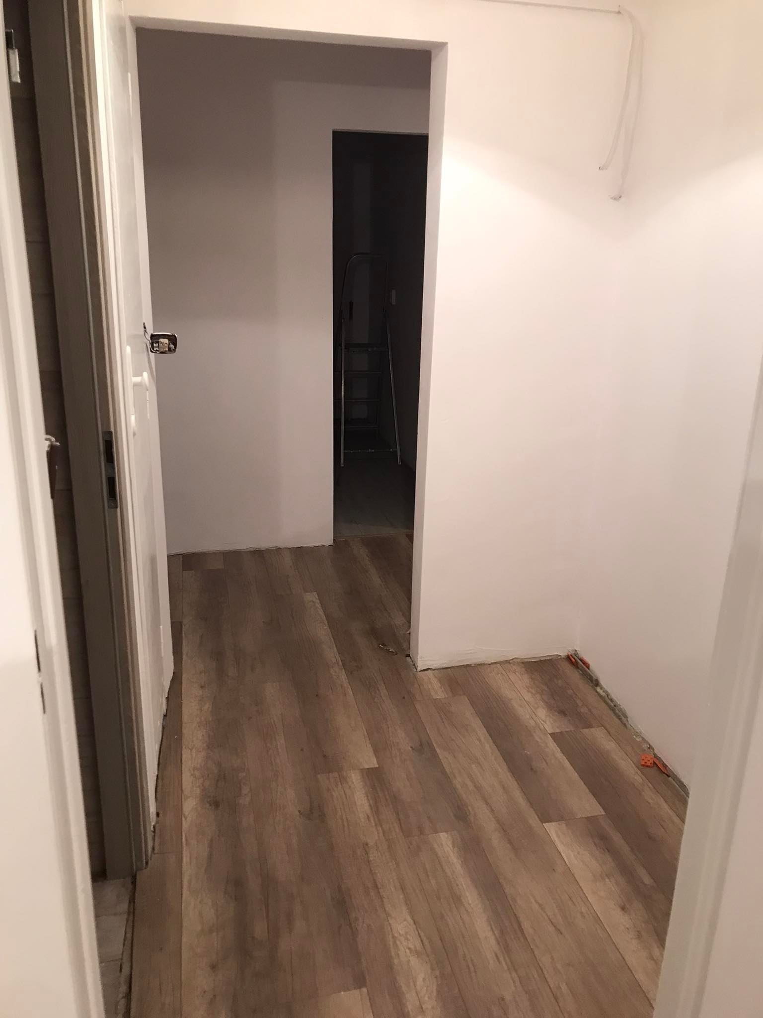 Korytarz po remoncie z panelami podłogowymi w odcieniach brązu, widoczne białe ściany i otwarte drzwi do kolejnego pomieszczenia z drabiną w tle.