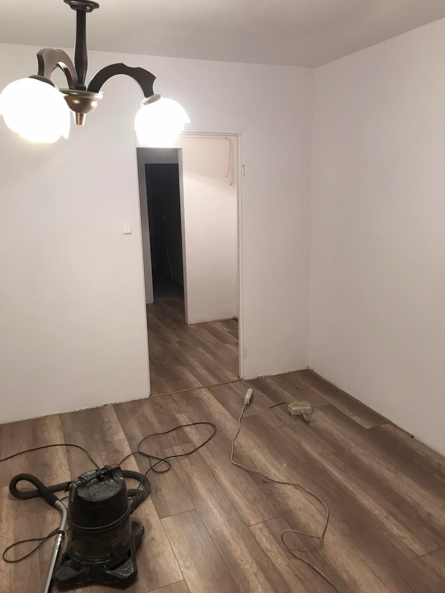 Pokój w trakcie układania paneli podłogowych, widoczny odkurzacz przemysłowy, kabel zasilający i szczotka do kleju.