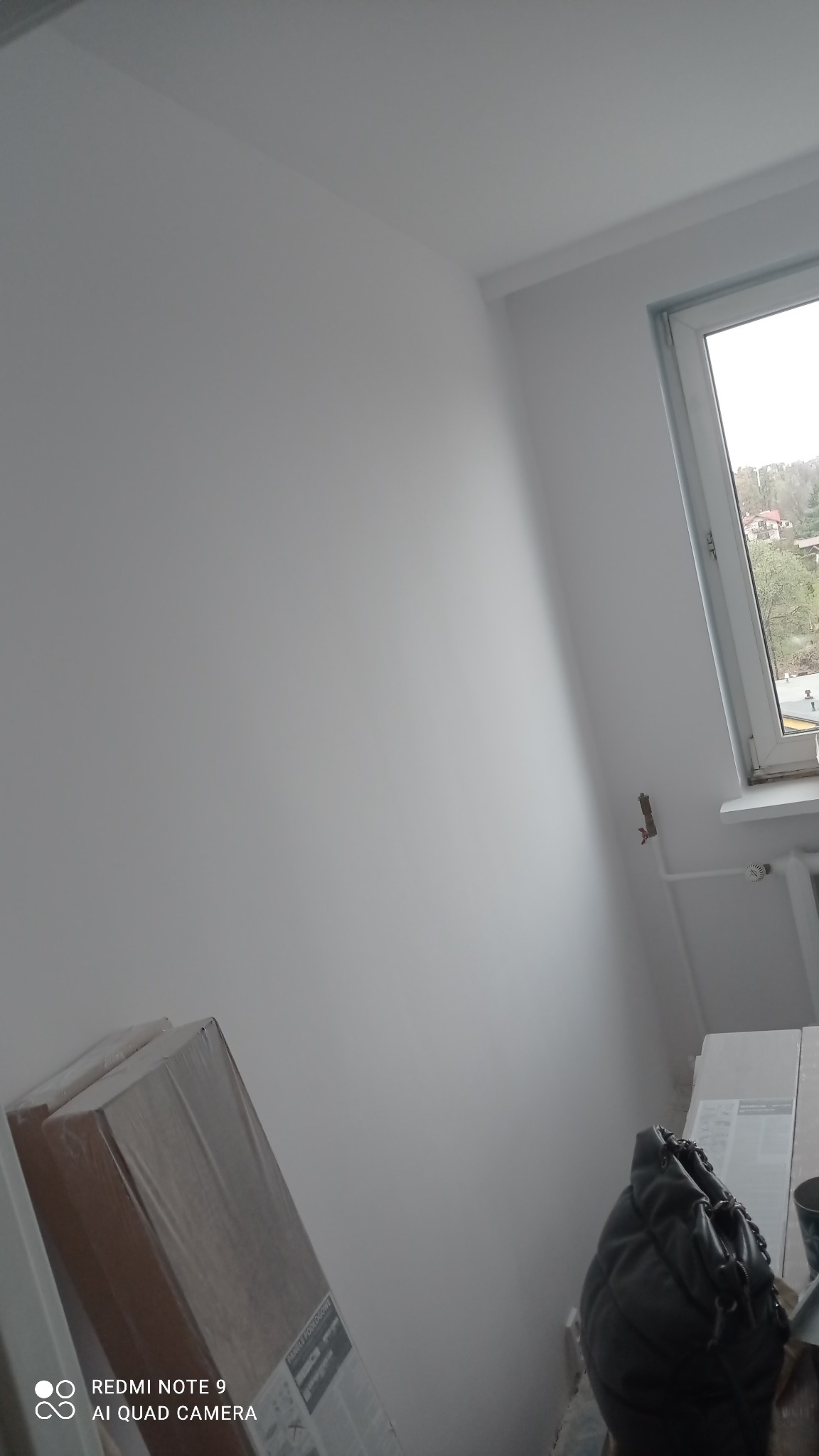 Świeżo pomalowane na biało wnętrze pokoju z oknem, widoczne zapakowane panele podłogowe i torba robocza.