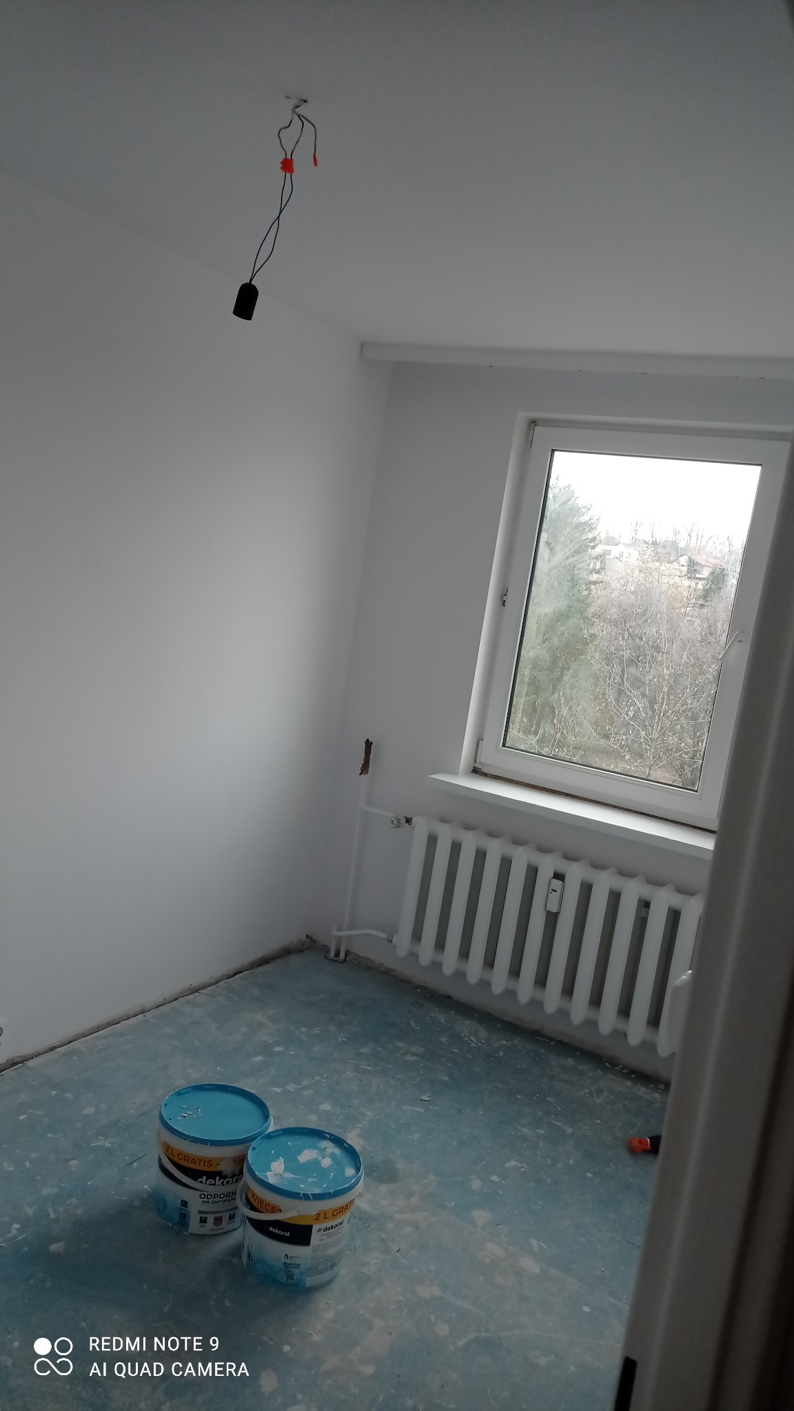 Pomalowane na biało wnętrze pokoju z dwoma wiaderkami farby Dekoral na podłodze zabezpieczonej folią, widoczne wiszące kable od oświetlenia i grzejnik pod oknem.