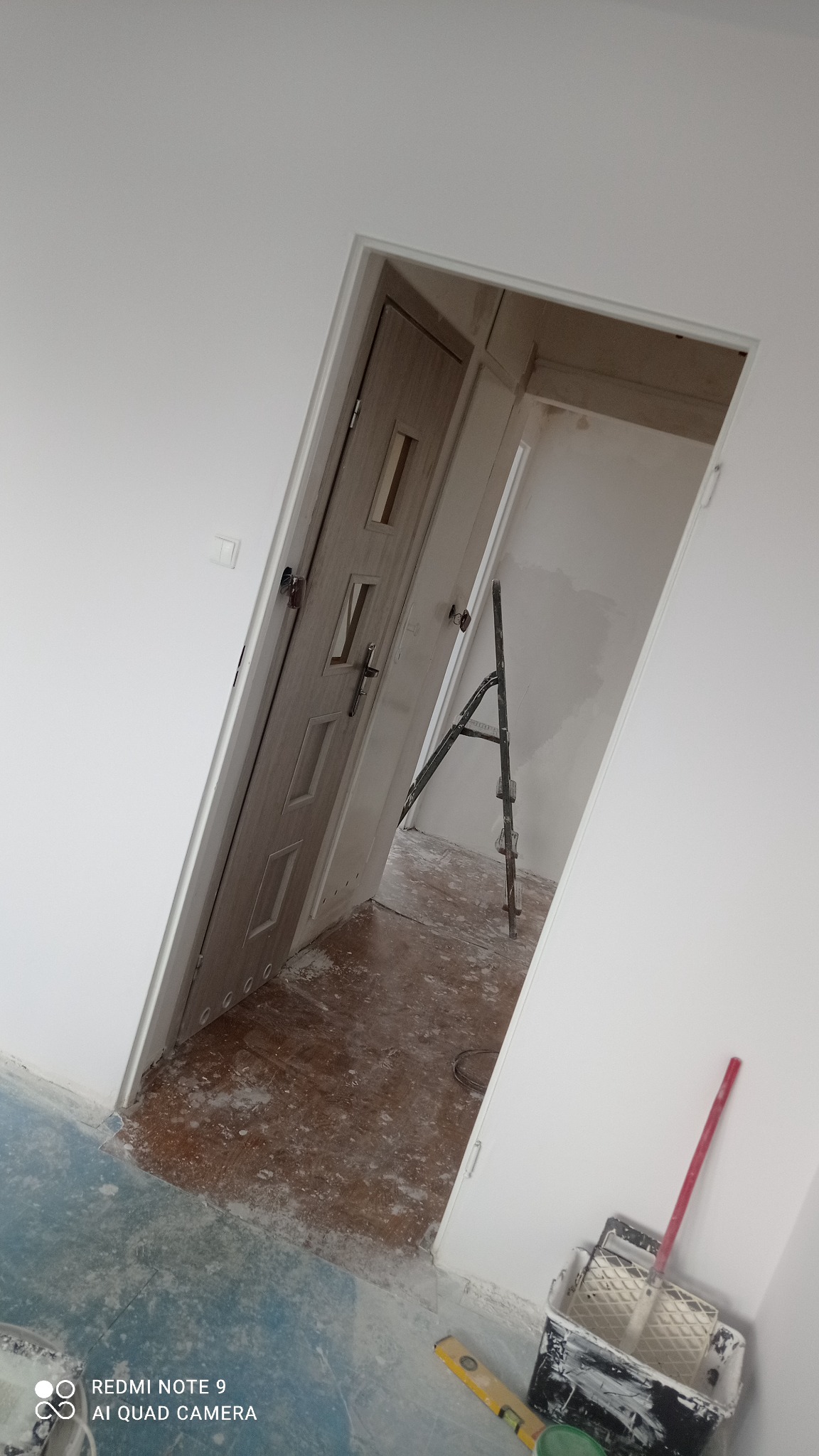 Wnętrze w trakcie malowania ścian na biało, widoczna drabina, drzwi wewnętrzne, poziomica i kuweta z wałkiem malarskim.