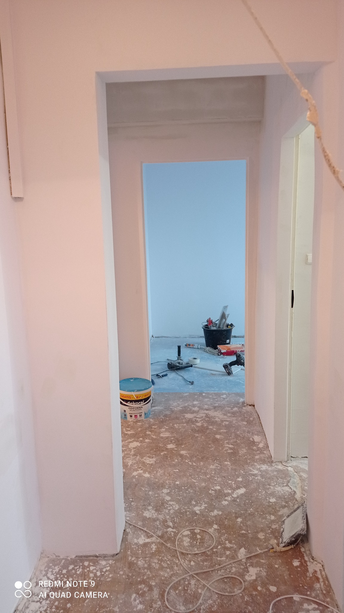 Korytarz w trakcie malowania na biało, widoczny kubeł z farbą Dekoral, narzędzia malarskie i zakurzona podłoga zabezpieczona folią malarską.