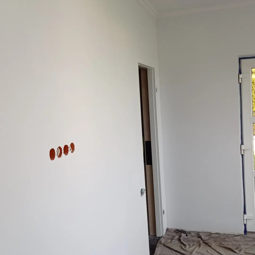 Biała ściana po obróbce, z czterema okrągłymi otworami pod gniazdka elektryczne, widoczny fragment drzwi i okna zabezpieczonego taśmą malarską.