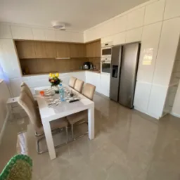 Jasna, nowoczesna kuchnia z białymi szafkami i drewnianymi akcentami, stół z krzesłami, bukiet tulipanów na blacie, widok z perspektywy podłogi z połyskliwymi płytkami.