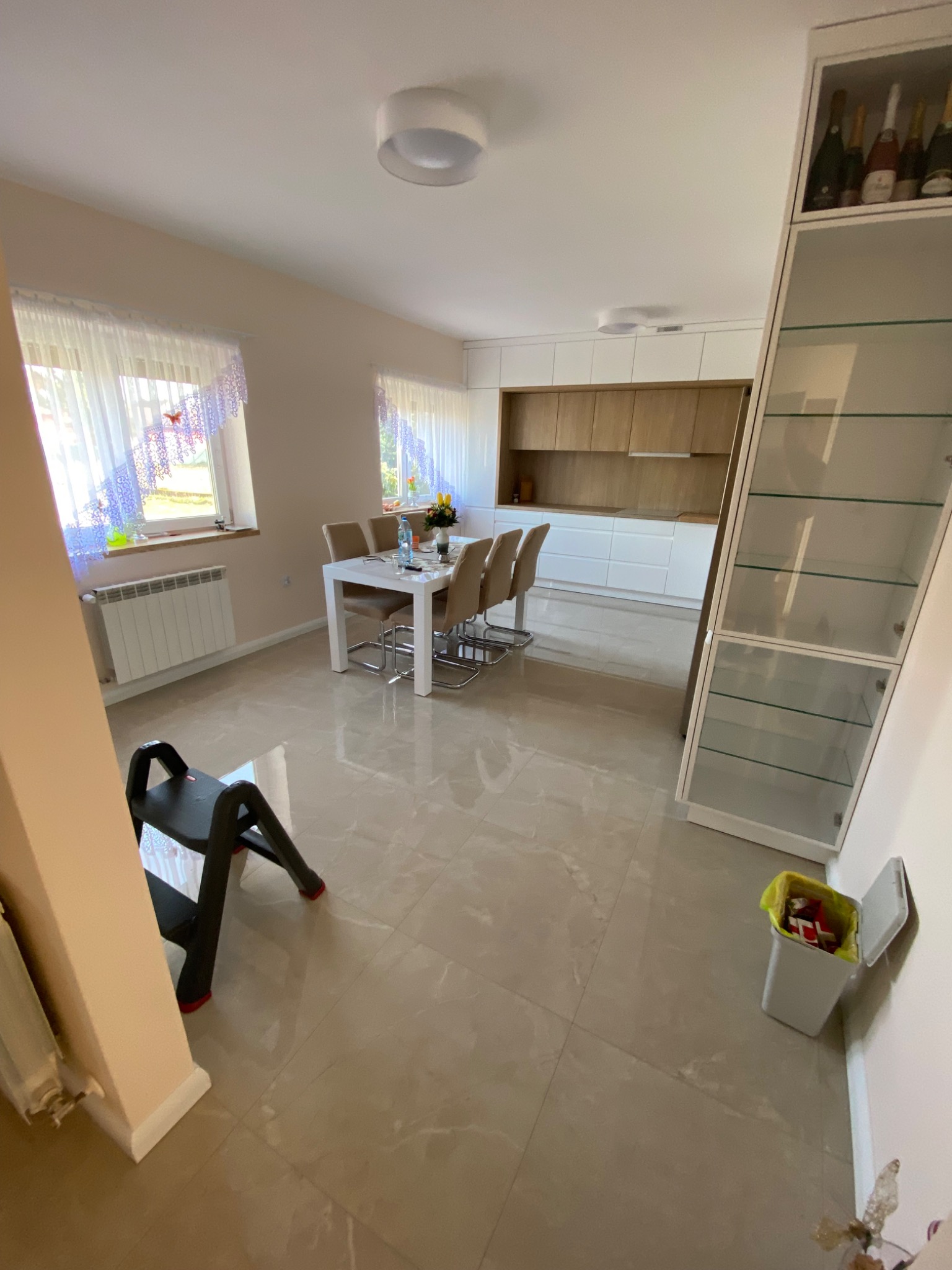 Jasne wnętrze po remoncie: płytki podłogowe z marmurkowym wzorem, biały stół z krzesłami, zabudowa kuchenna z drewnianymi frontami, szklana witryna, drabinka malarska i kosz na śmieci.