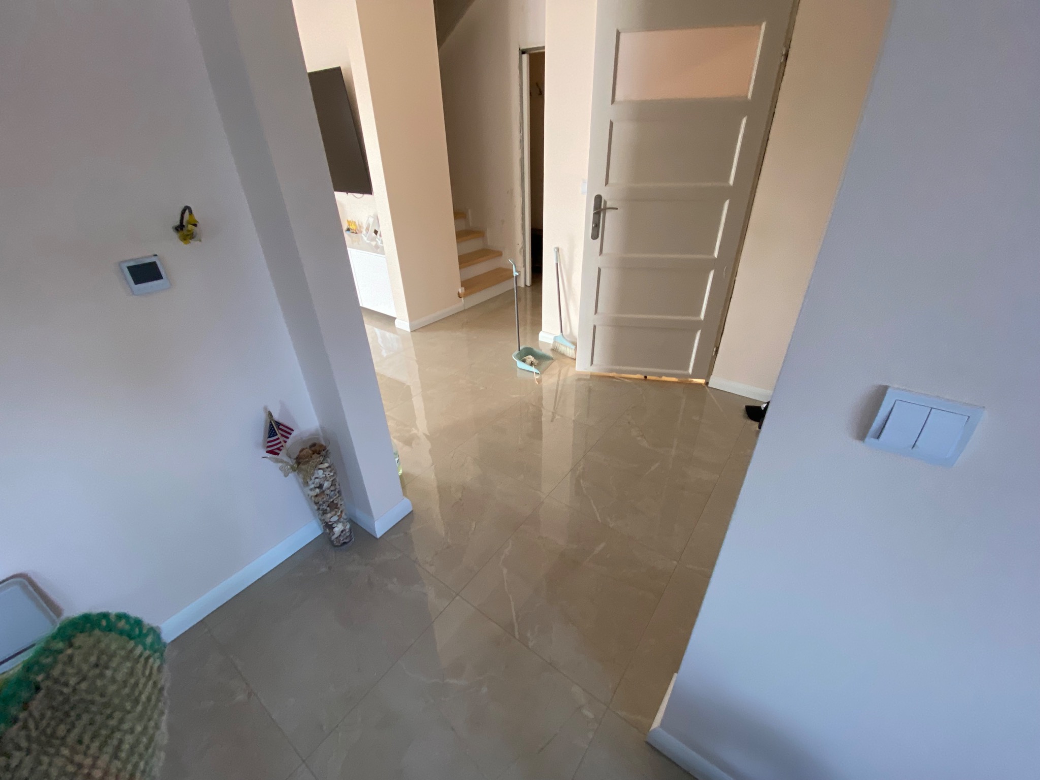 Beżowe płytki podłogowe z połyskiem w przedpokoju, widoczny mop i zmiotka, białe ściany i drzwi, w tle schody.