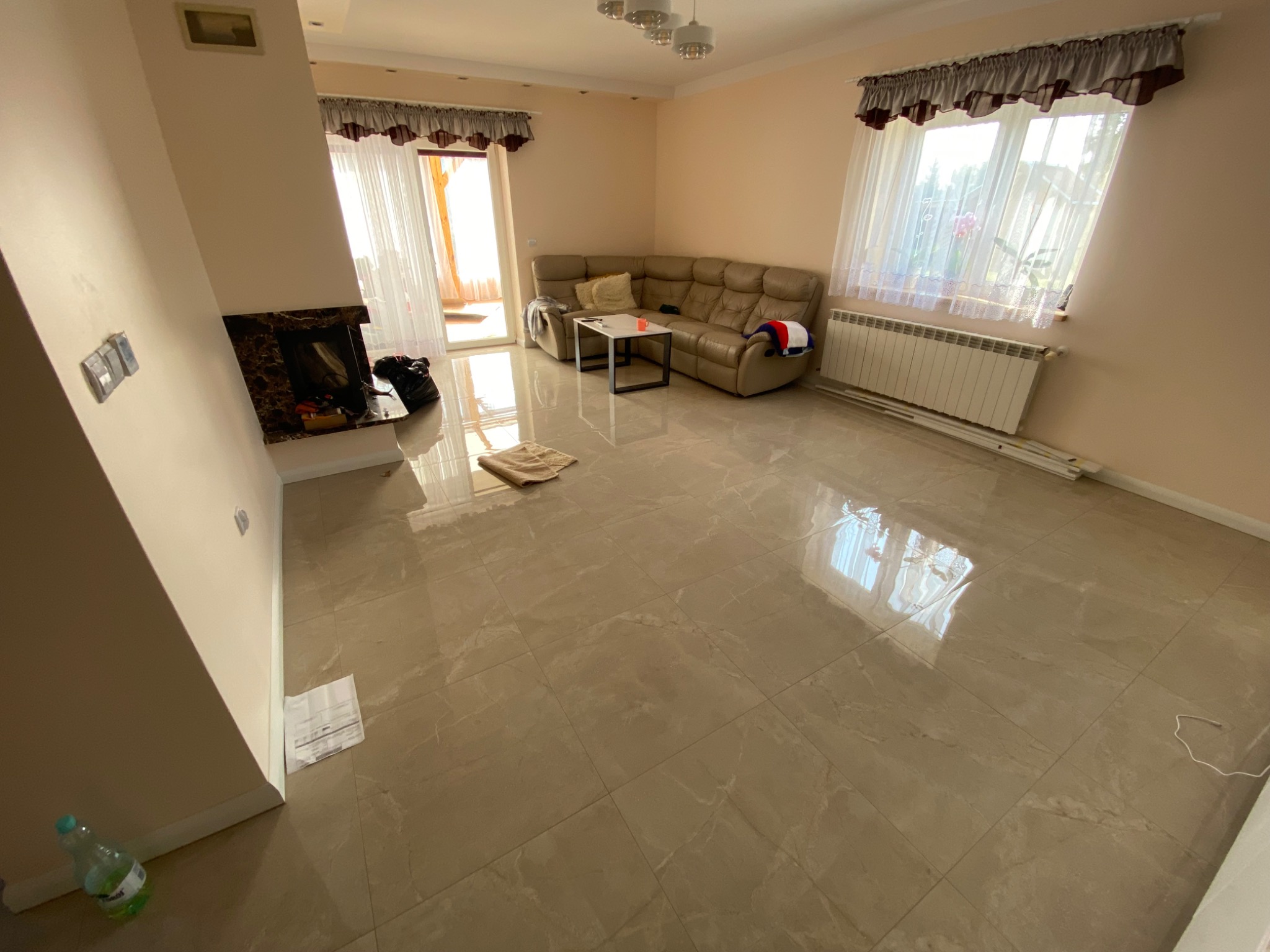 Salon po remoncie: beżowe płytki podłogowe z połyskiem, jasne ściany, narożnik i kominek z ciemnym marmurem, widoczne niedoskonałości wykończenia.