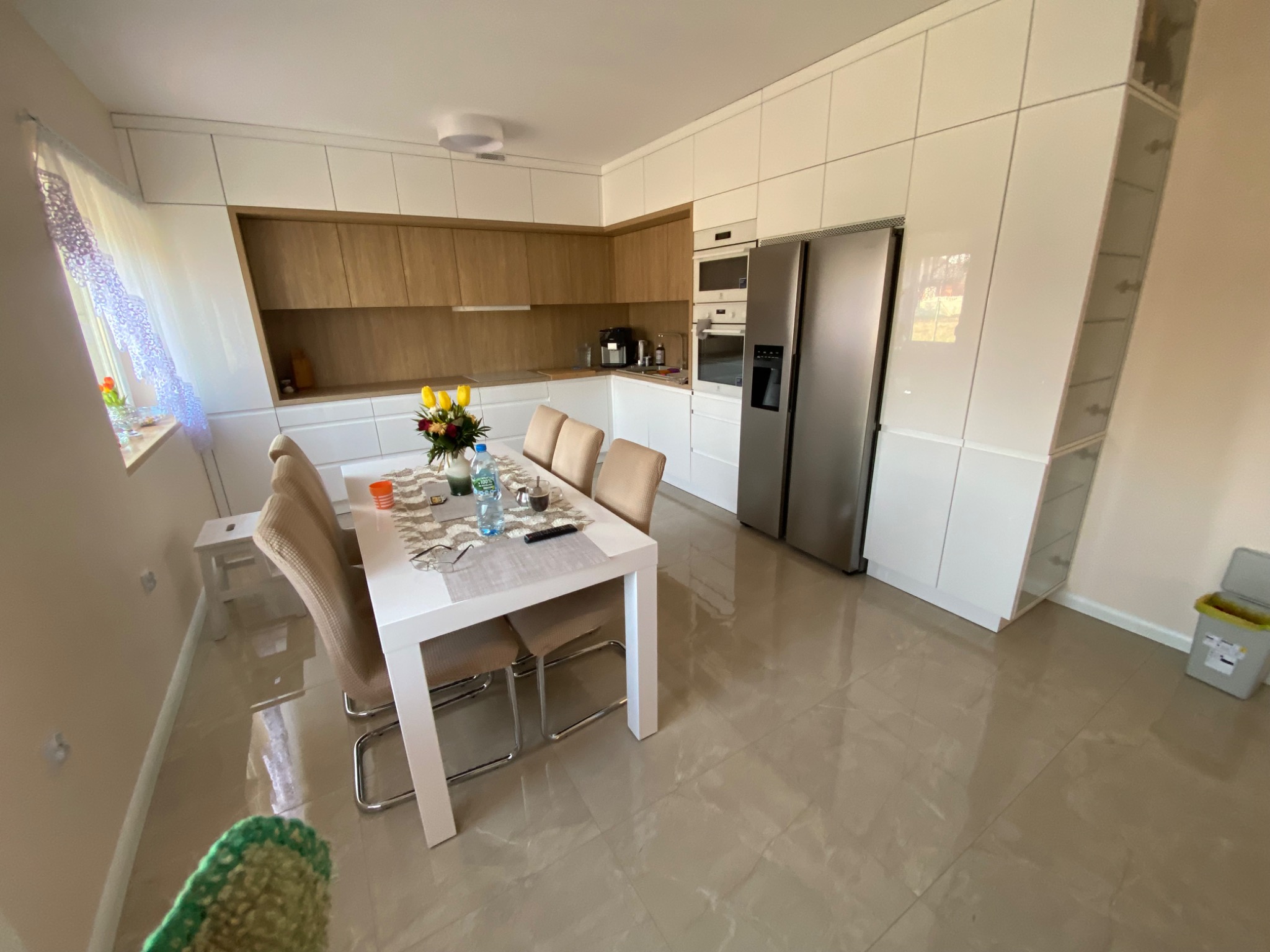 Jasna, nowoczesna kuchnia z białymi szafkami i drewnianymi akcentami, stół z krzesłami, bukiet tulipanów na blacie, widok z perspektywy podłogi z połyskliwymi płytkami.