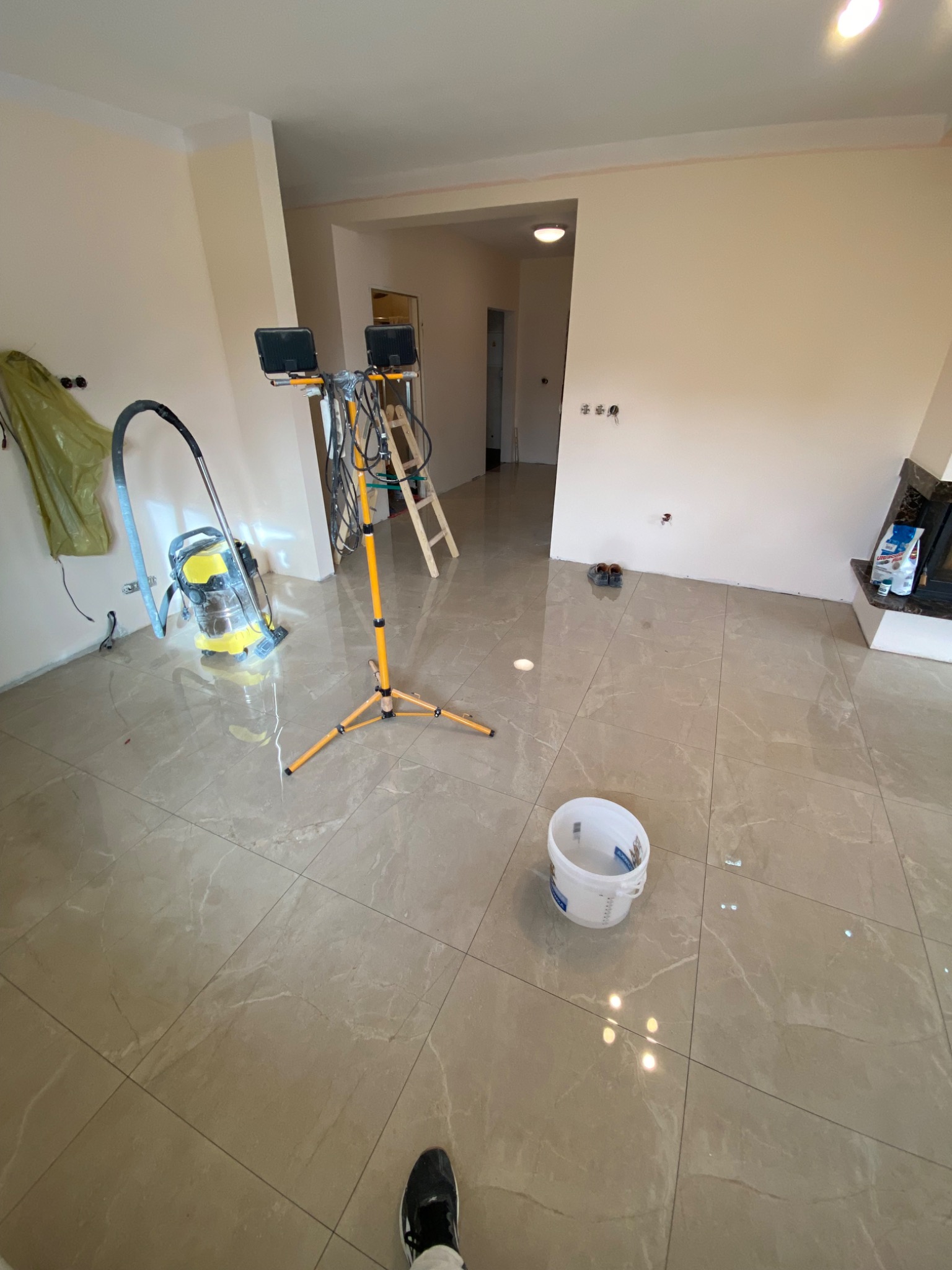 Salon w trakcie prac wykończeniowych z nową, błyszczącą podłogą z płytek, widoczny odkurzacz przemysłowy, wiadro z farbą i lampa budowlana na trójnogu.
