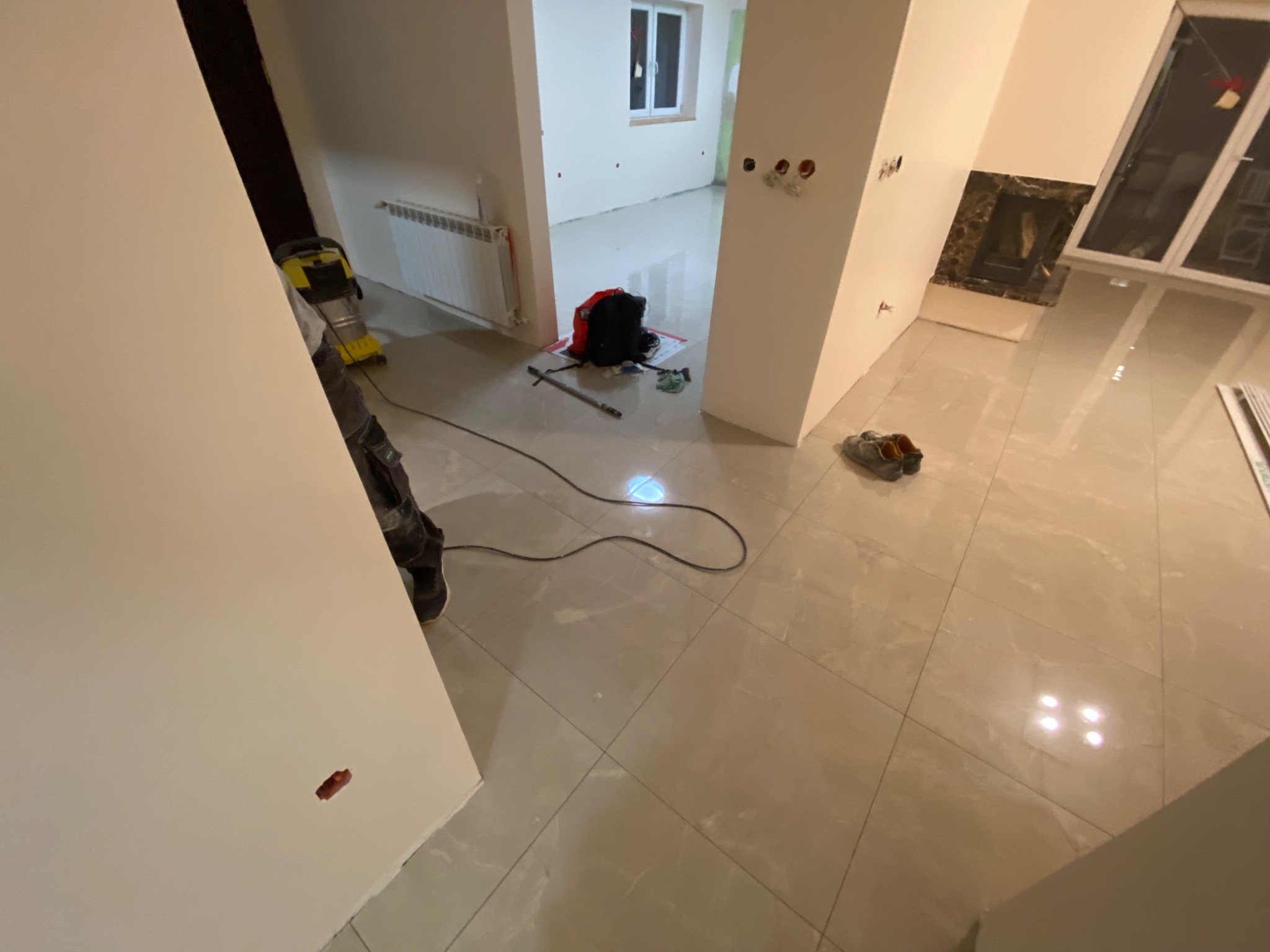 Wnętrze w trakcie wykańczania: świeżo położone, błyszczące płytki podłogowe, widoczny sprzęt budowlany, grzejnik oraz przygotowane punkty elektryczne na ścianie.