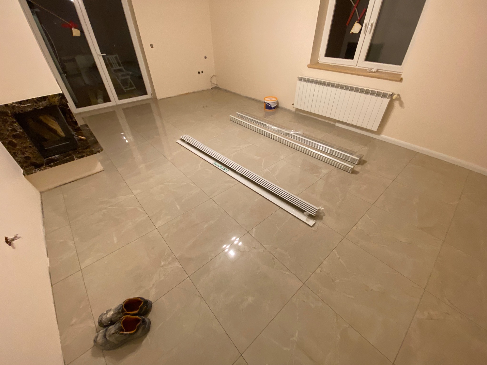 Pokój w trakcie remontu: ułożone płytki podłogowe, przygotowane listwy przypodłogowe, widoczny kominek z obudową z ciemnego marmuru, grzejnik pod oknem, na podłodze robocze buty.