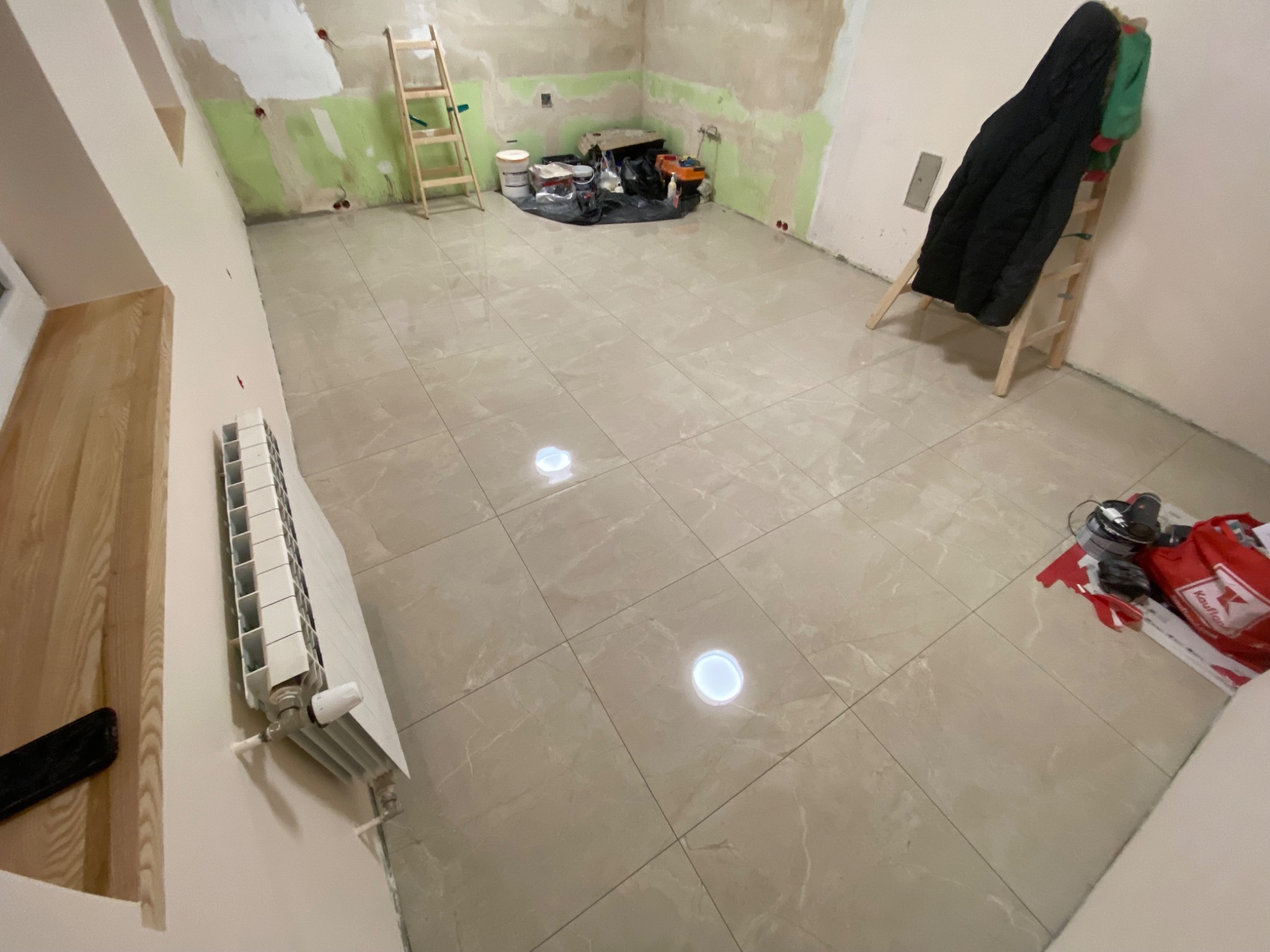 Ułożone beżowe płytki podłogowe z marmurkowym wzorem w pokoju w trakcie remontu; widoczny grzejnik, drabina oparta o ścianę, narzędzia i materiały budowlane.