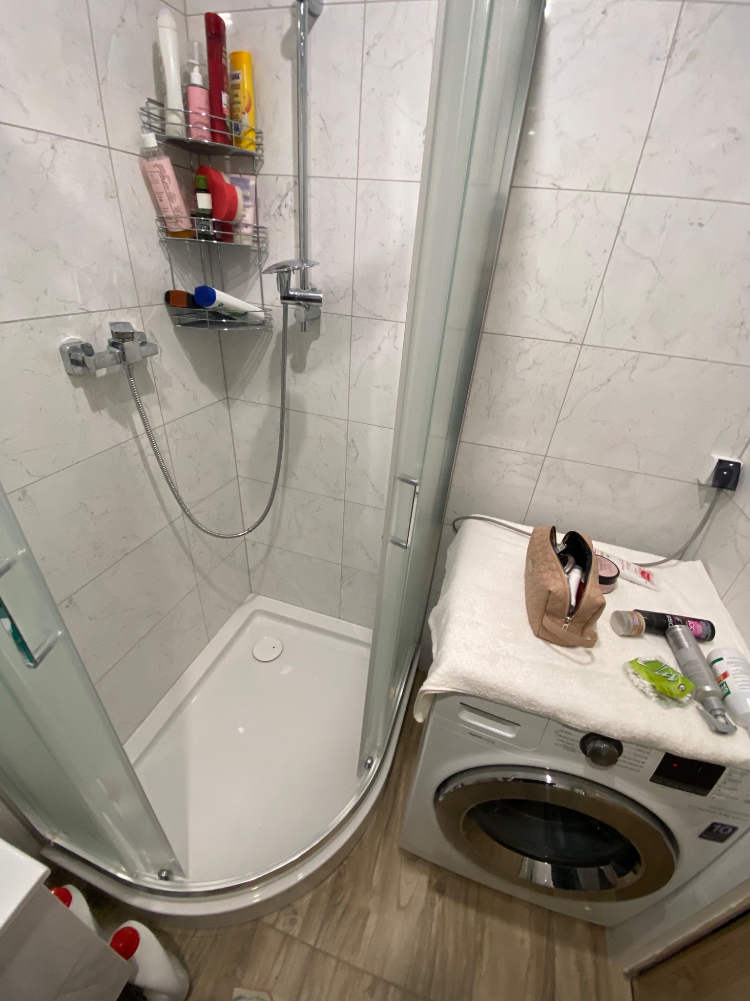 Ujęcie narożnej kabiny prysznicowej z brodzikiem, widoczne półki na kosmetyki, bateria prysznicowa oraz pralka z kosmetykami na wierzchu, wykończona płytkami imitującymi marmur.