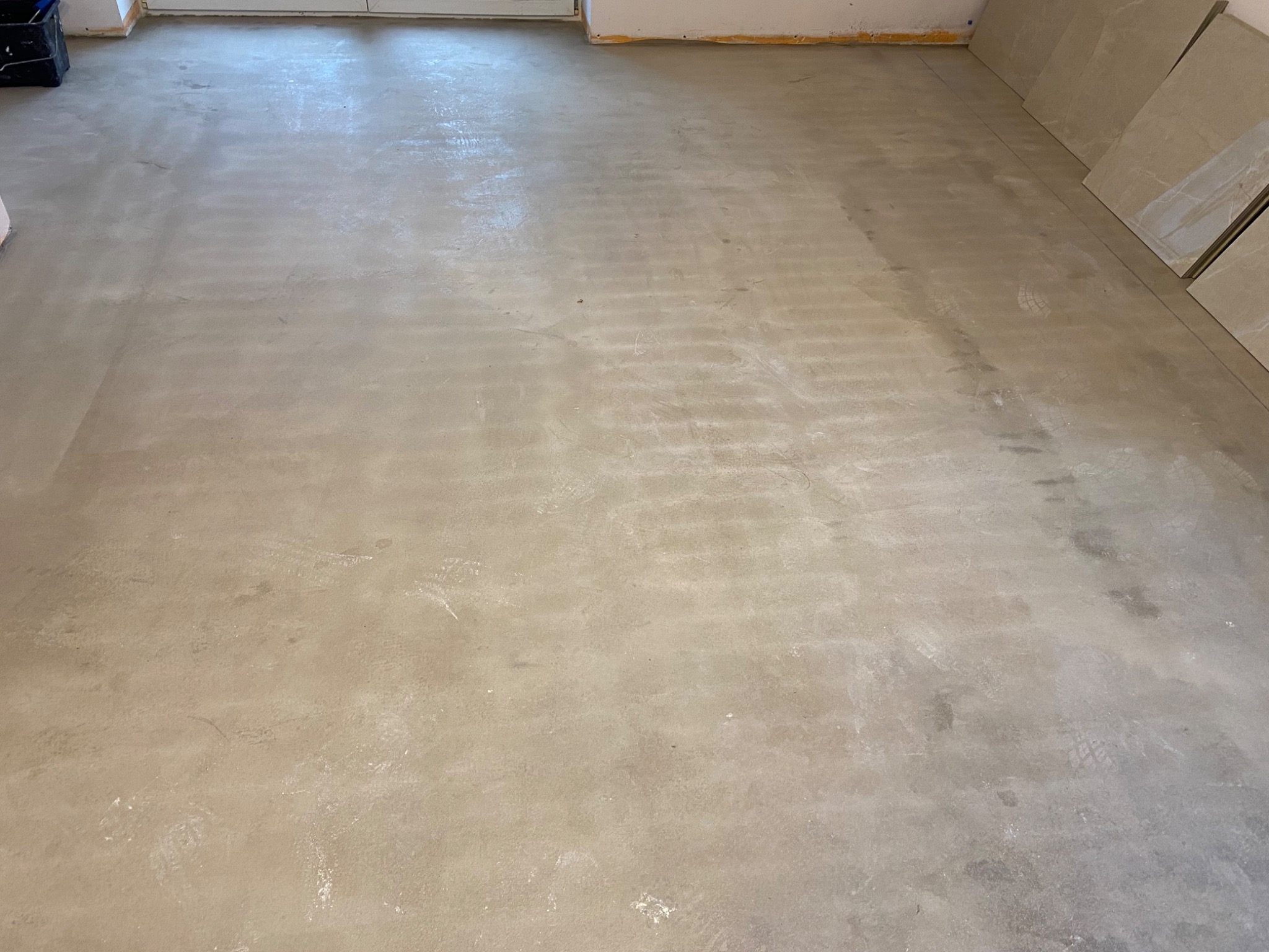 Przygotowane podłoże pod płytki, z widocznymi śladami stóp i ułożonymi płytkami w rogu pomieszczenia.