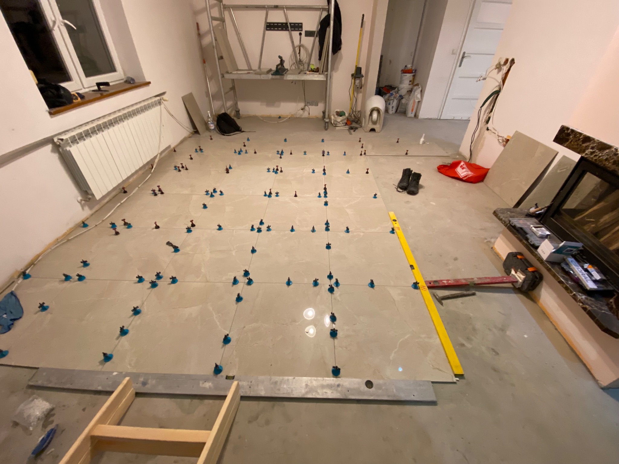 Układanie płytek podłogowych z użyciem systemu poziomowania, widoczne krzyżyki dystansowe i poziomica, drabina w tle.