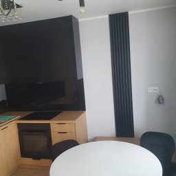 Apo-Inwestment Artur Popis - Nowoczesna kuchnia z zabudową w odcieniach beżu i czerni, okrągły biały stół z czarnymi krzesłami, dekoracyjne czarne lamele na ścianie, minimalistyczne oświetlenie.