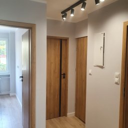 Apo-Inwestment Artur Popis - Korytarz po odświeżeniu: szare ściany, drewniane drzwi z czarnymi akcentami, jasna podłoga imitująca drewno, oświetlenie punktowe na szynie.