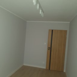 Apo-Inwestment Artur Popis - Pusty, szary pokój z drewnianą podłogą i drzwiami z ciemnym paskiem, oświetlony reflektorami sufitowymi