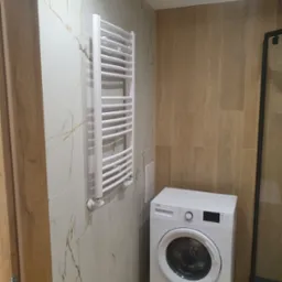 Wykończona łazienka z białą pralką Beko, grzejnikiem drabinkowym na ścianie z płytkami imitującymi marmur oraz ścianą wyłożoną panelami drewnopodobnymi.