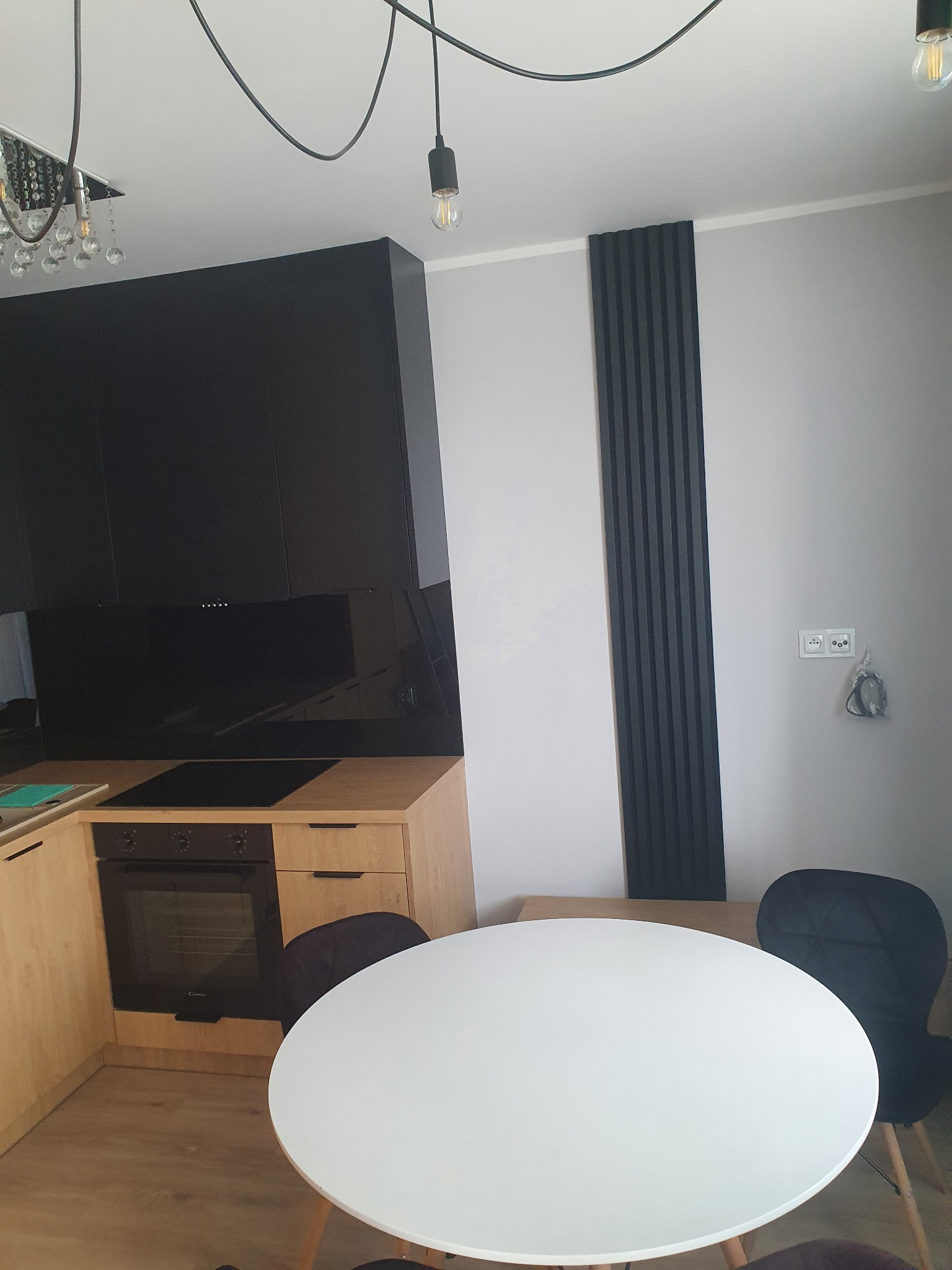 Nowoczesna kuchnia z zabudową w odcieniach beżu i czerni, okrągły biały stół z czarnymi krzesłami, dekoracyjne czarne lamele na ścianie, minimalistyczne oświetlenie.