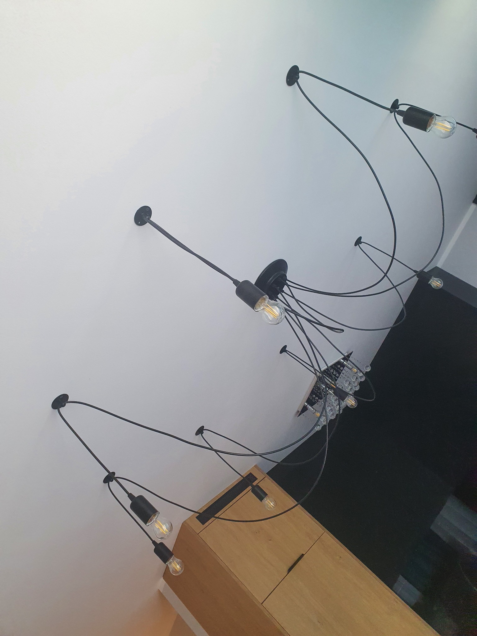 Nowoczesna lampa sufitowa z kilkoma czarnymi przewodami i żarówkami Edisona, zamontowana na białym suficie, widoczna z perspektywy żabiej, z fragmentem drewnianej komody w tle.