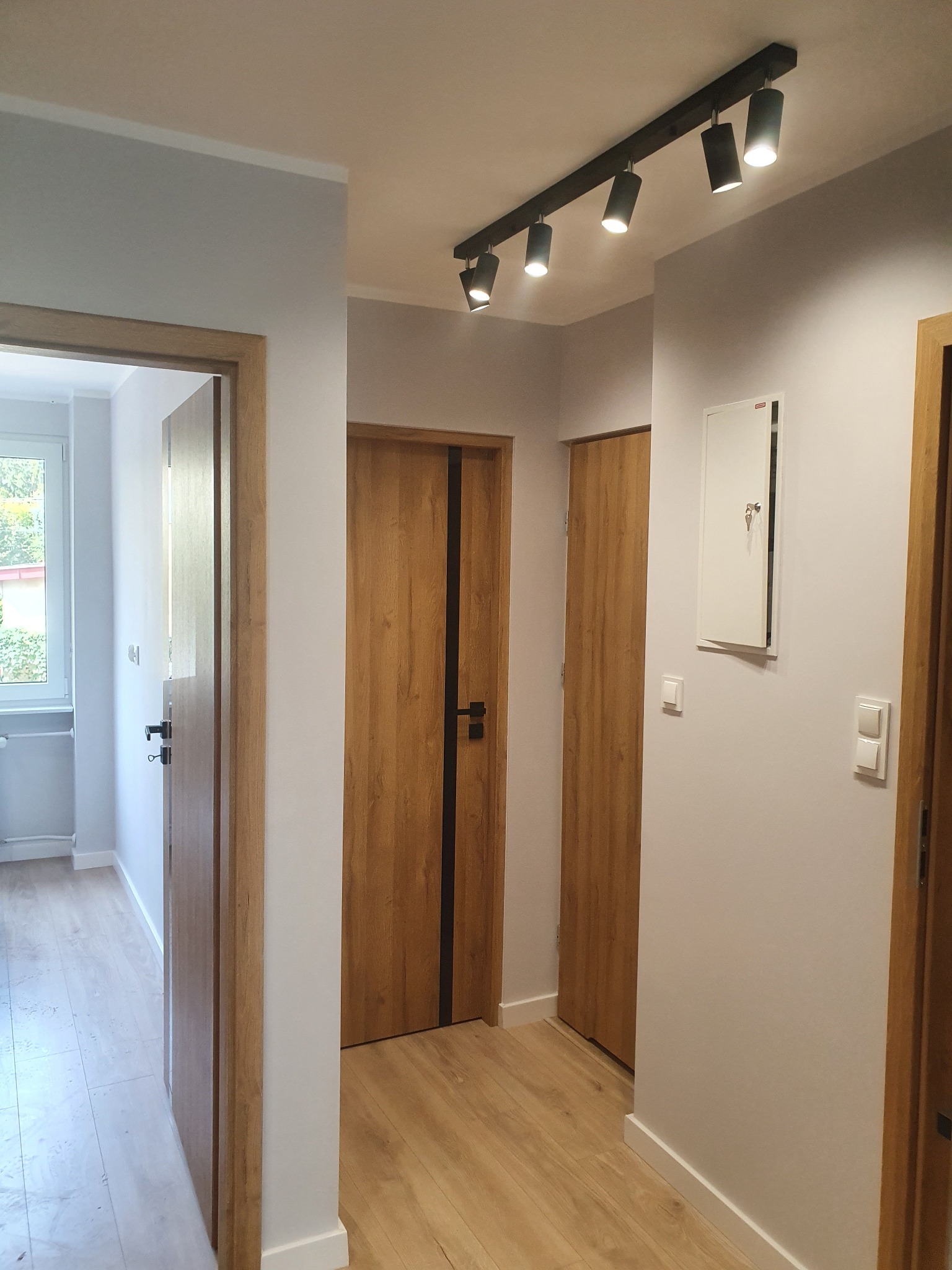 Korytarz po odświeżeniu: szare ściany, drewniane drzwi z czarnymi akcentami, jasna podłoga imitująca drewno, oświetlenie punktowe na szynie.