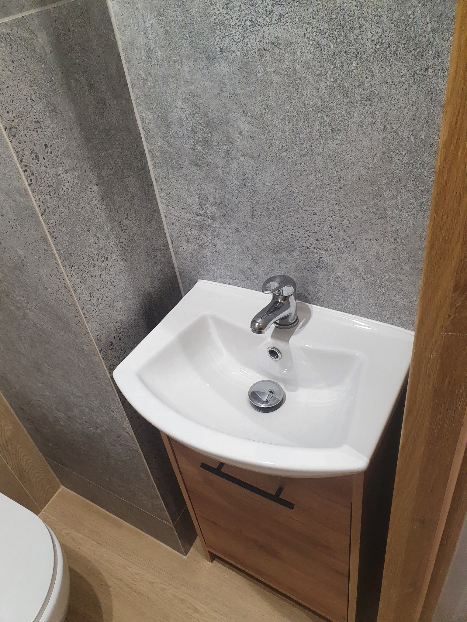Umywalka nablatowa z szafką w łazience wykończonej szarą płytką imitującą kamień, widoczny fragment toalety i drewniany element wykończeniowy.