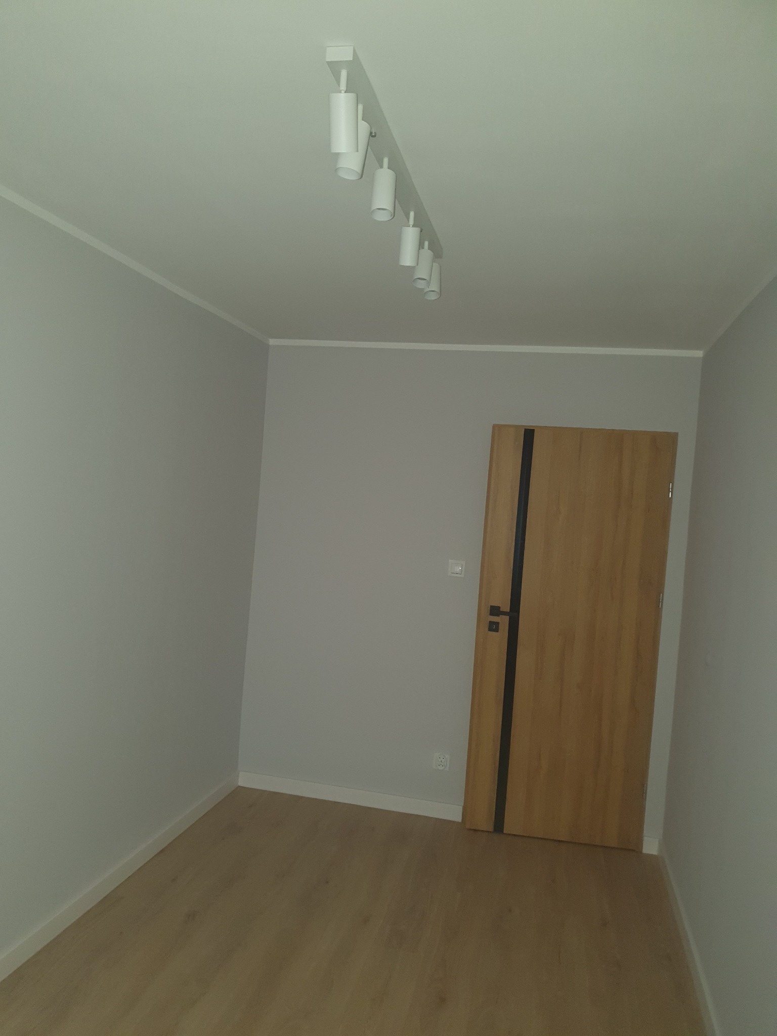 Pusty, szary pokój z drewnianą podłogą i drzwiami z ciemnym paskiem, oświetlony reflektorami sufitowymi