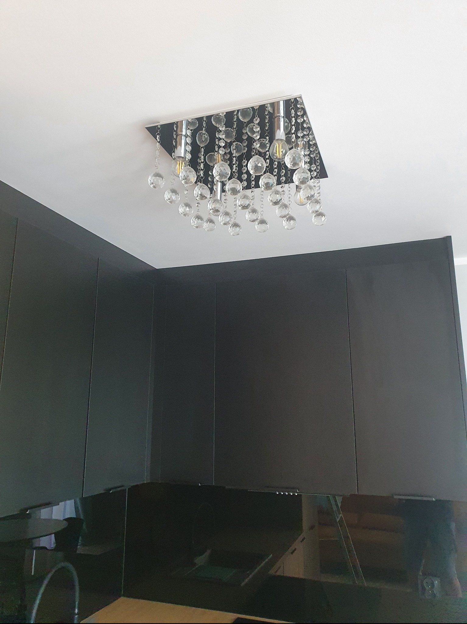 Nowoczesna lampa sufitowa z kryształkami w kwadratowej oprawie, zamontowana w ciemnej kuchni z błyszczącymi szafkami.