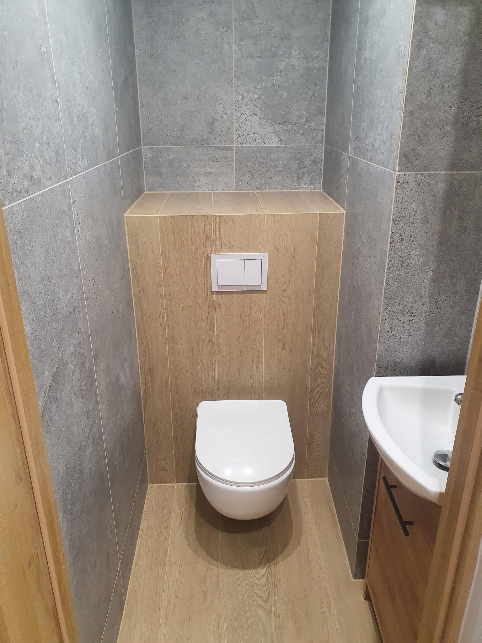 Wnętrze małej toalety z szarą płytką imitującą kamień na ścianach, drewnopodobną okładziną na podłodze i zabudowie stelaża podtynkowego, wisząca muszla klozetowa i umywalka nablatowa z szafką.