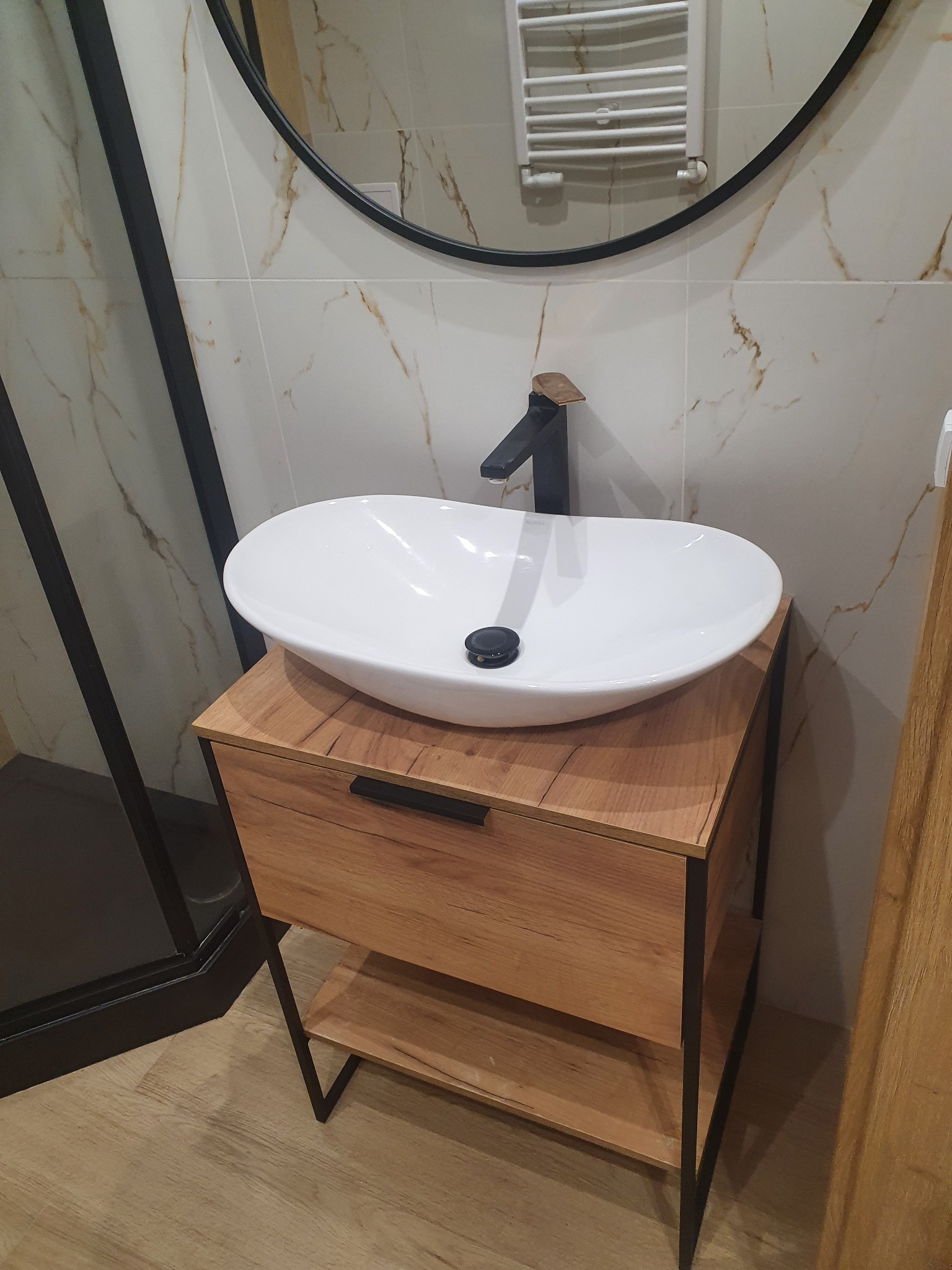 Nowoczesna łazienka: biała umywalka nablatowa na szafce z drewnianym frontem i czarnymi metalowymi nogami, czarna bateria, okrągłe lustro z czarną ramą, płytki imitujące marmur.