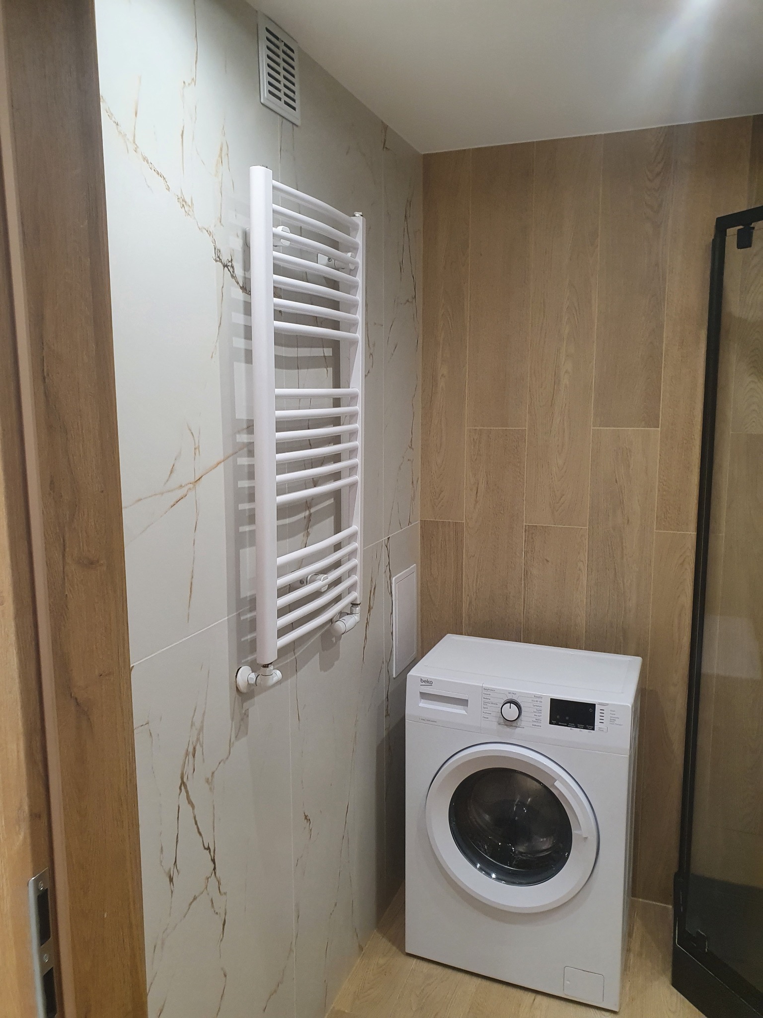 Wykończona łazienka z białą pralką Beko, grzejnikiem drabinkowym na ścianie z płytkami imitującymi marmur oraz ścianą wyłożoną panelami drewnopodobnymi.