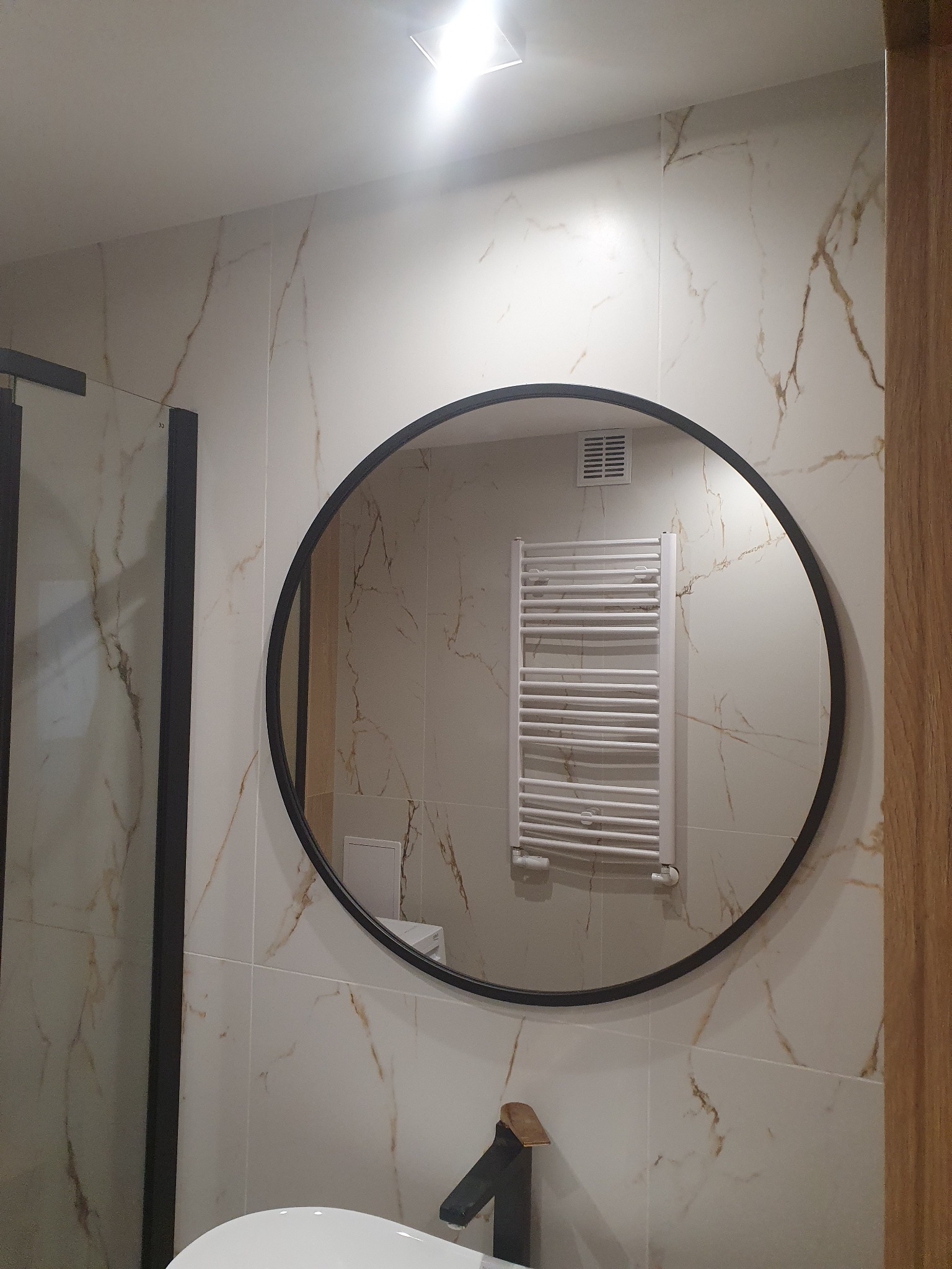 Okrągłe lustro w czarnej ramie odbija grzejnik drabinkowy i białe płytki imitujące marmur w łazience z czarną armaturą.