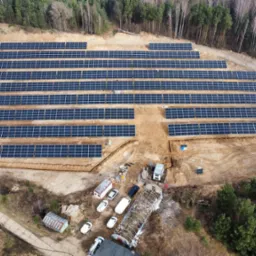 Widok z lotu ptaka na farmę fotowoltaiczną w Chmielowie o mocy 1MW, z panelami ułożonymi rzędami na przygotowanym terenie, widoczne zaplecze budowy z kontenerami i pojazdami.