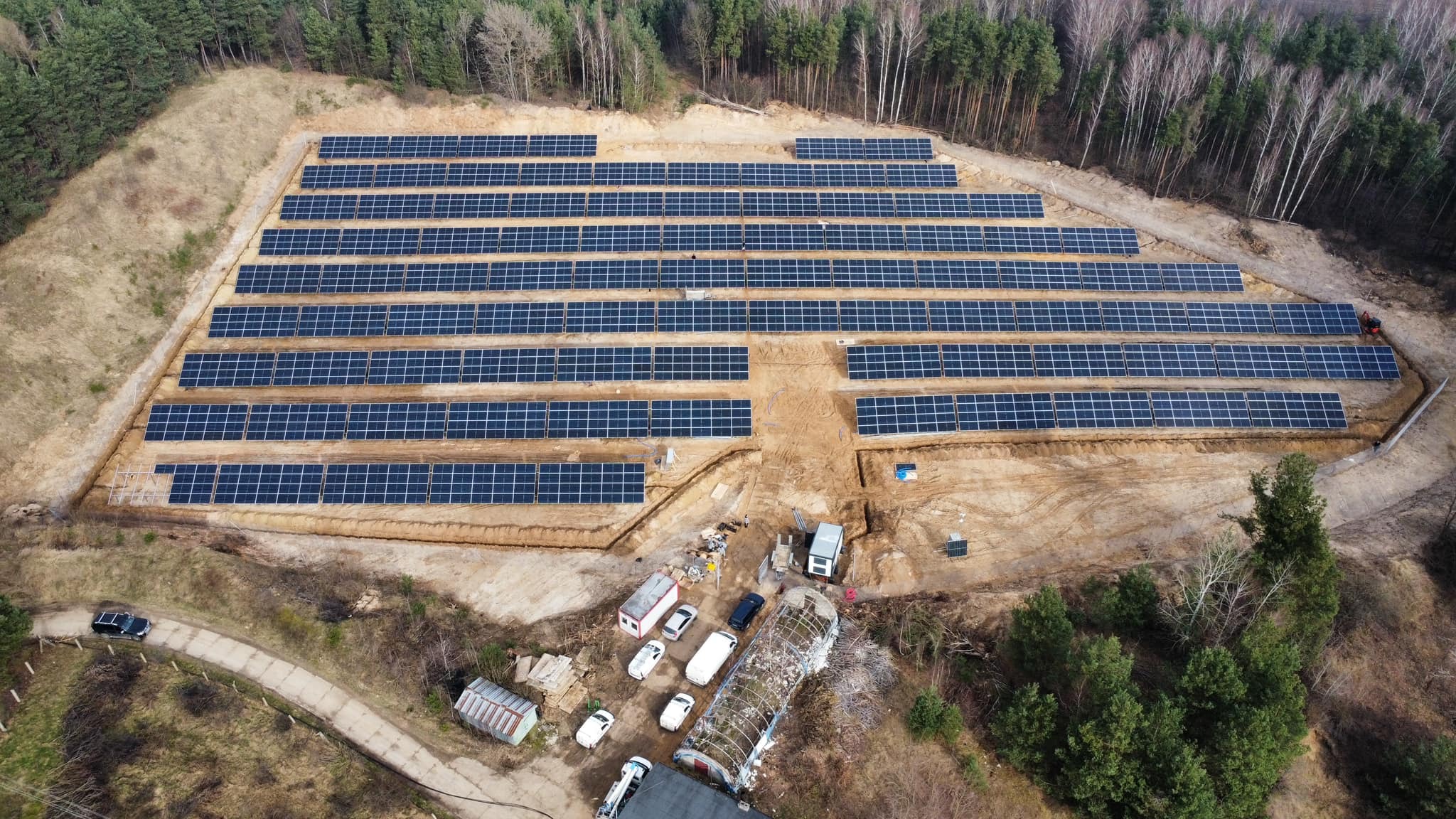 Widok z lotu ptaka na farmę fotowoltaiczną w Chmielowie o mocy 1MW, z panelami ułożonymi rzędami na przygotowanym terenie, widoczne zaplecze budowy z kontenerami i pojazdami.