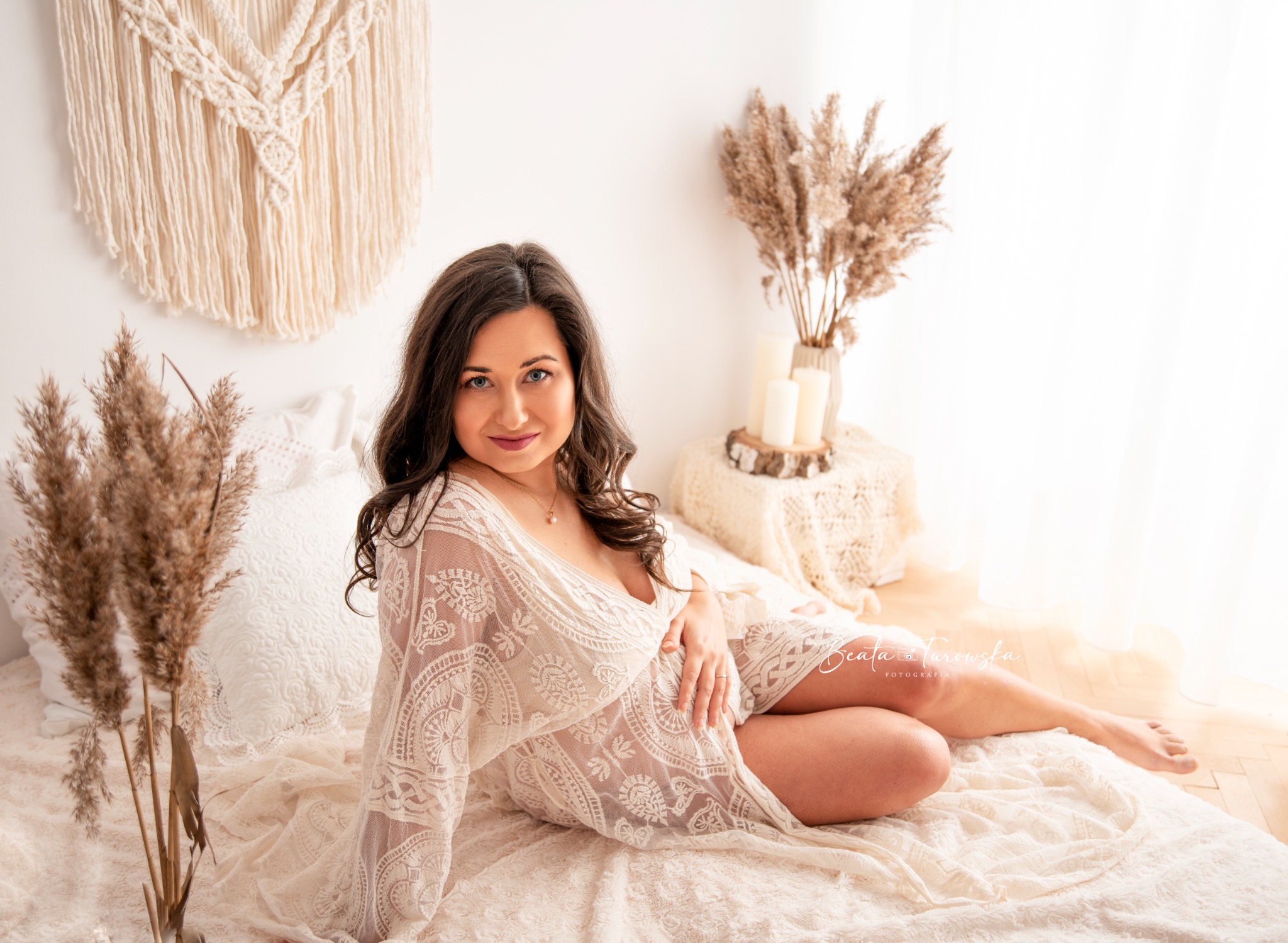 Kobieta w ciąży w koronkowej sukience pozuje na łóżku z dekoracjami w stylu boho, w tle makrama i suszone trawy.